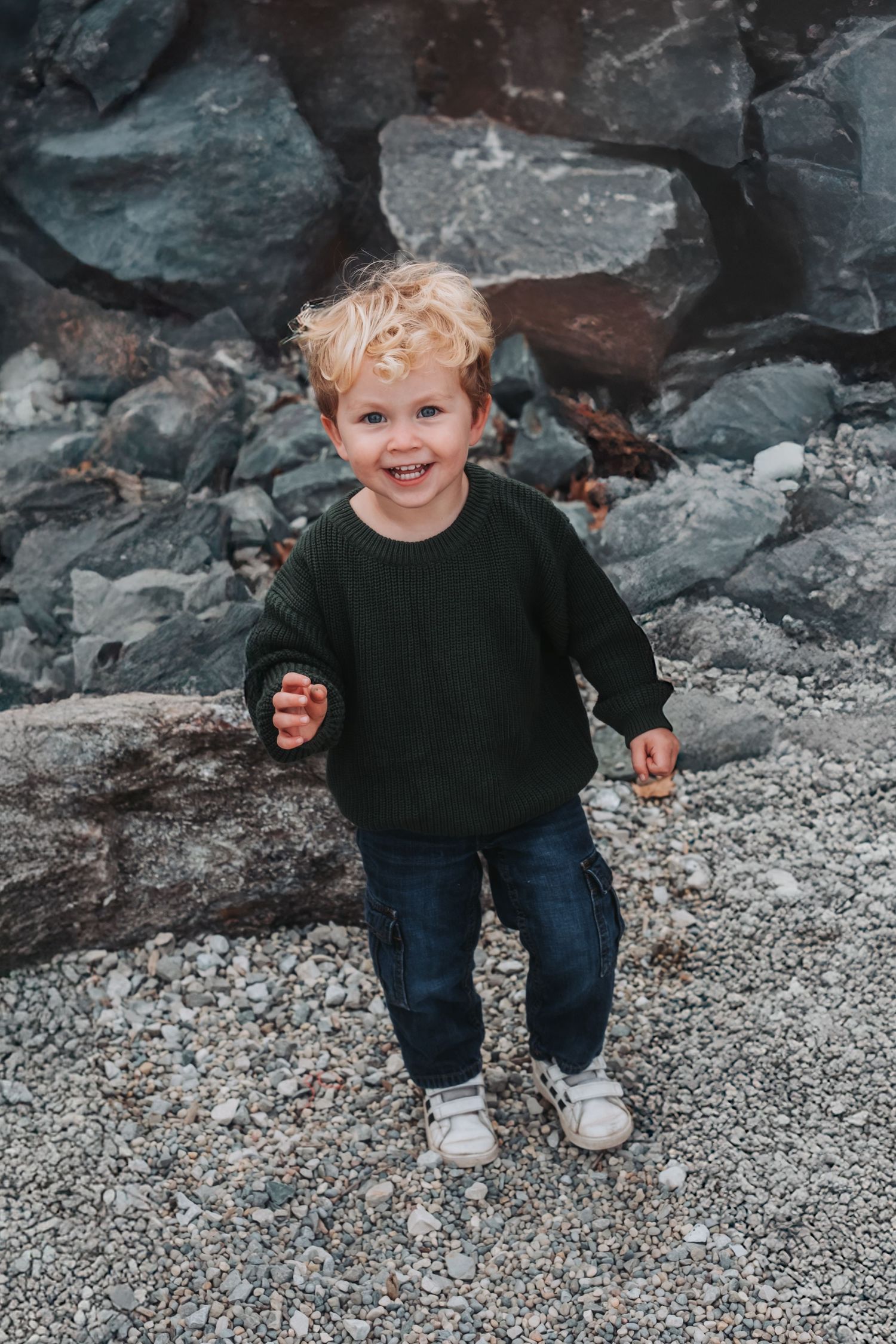 Small child sitting on rocky ground wearing dark clothing and white shoes.