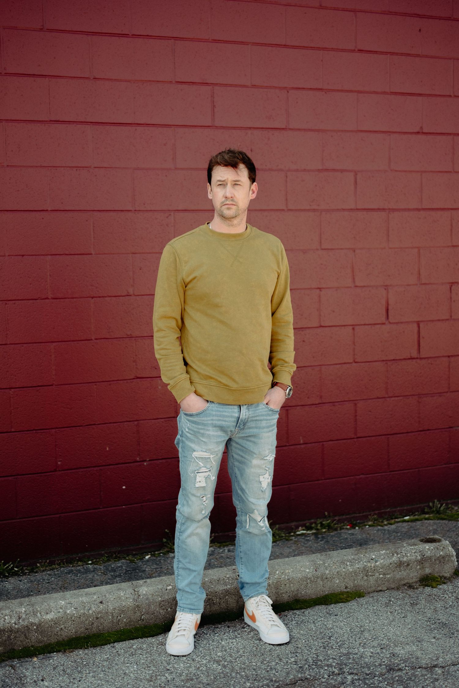 Person in yellow sweater and ripped jeans standing against red brick wall.