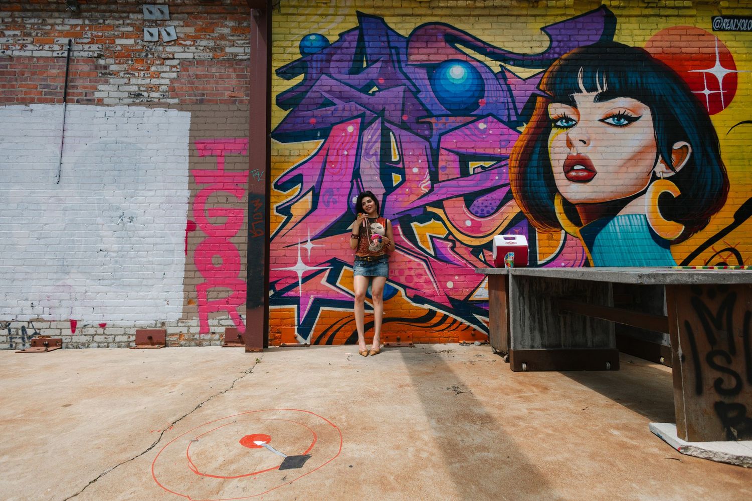 Woman posing in front of colorful graffiti mural featuring bold lettering and portrait art on brick wall.