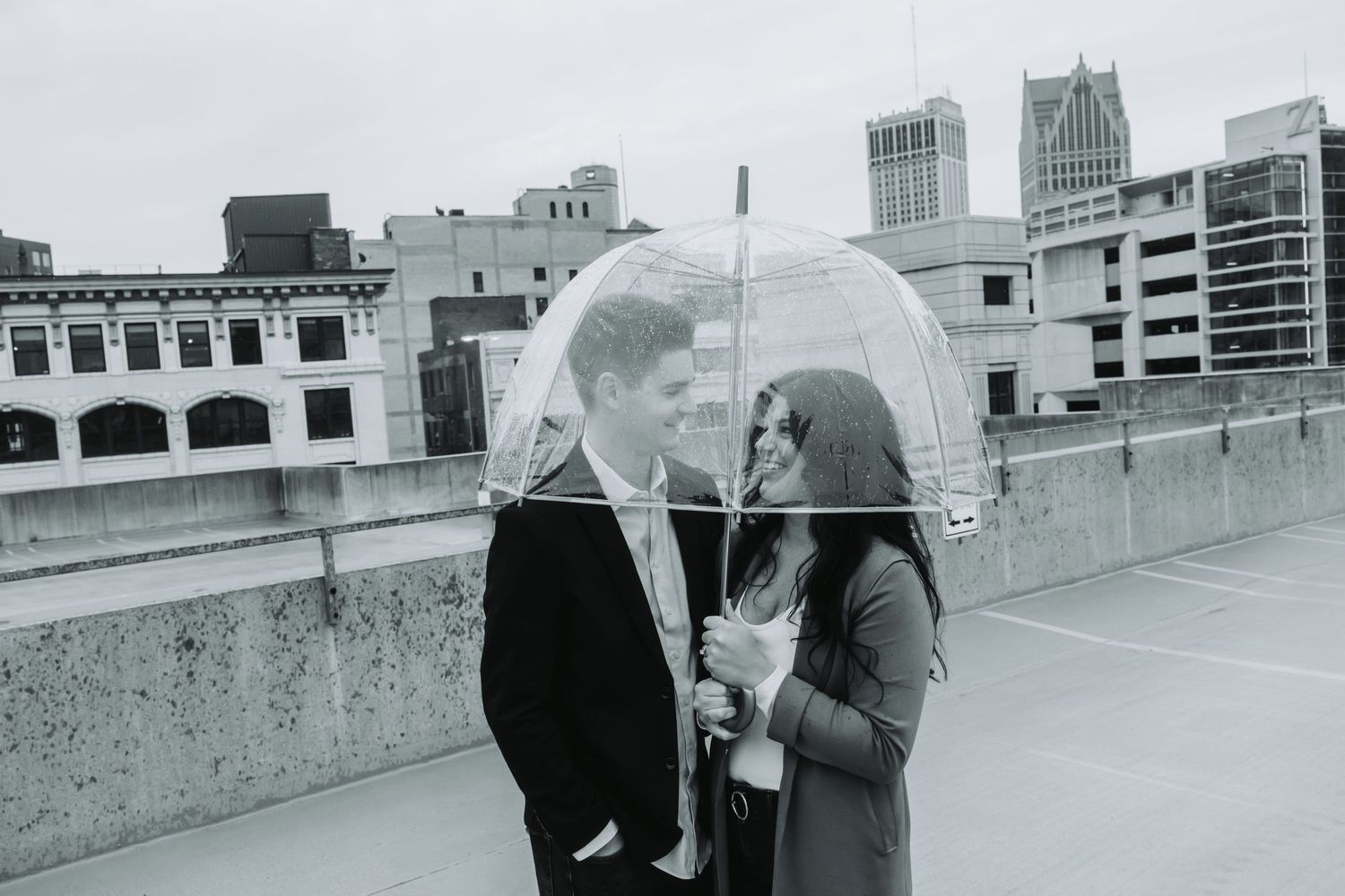 Couple sharing a clear umbrella on a rooftop with city skyline in black and white photo.