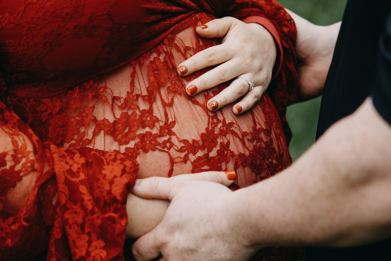 Couple tenderly holding pregnant belly in red lace maternity dress with matching red nail polish and ring.