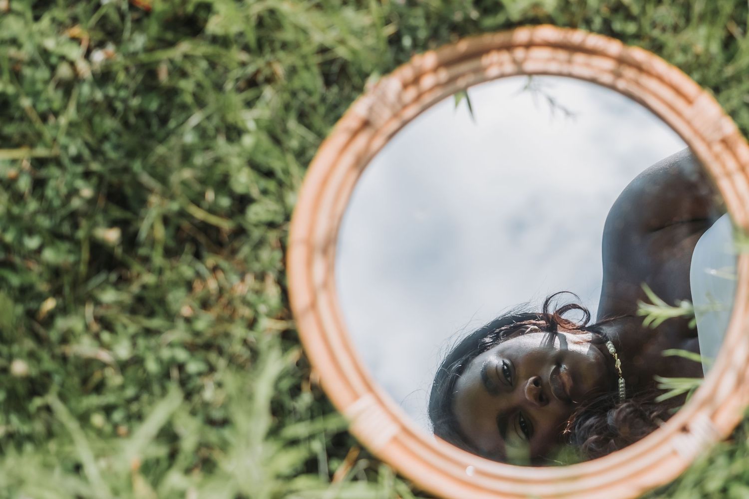 A young woman with braids reflects in a round mirror lying on green grass outdoors.