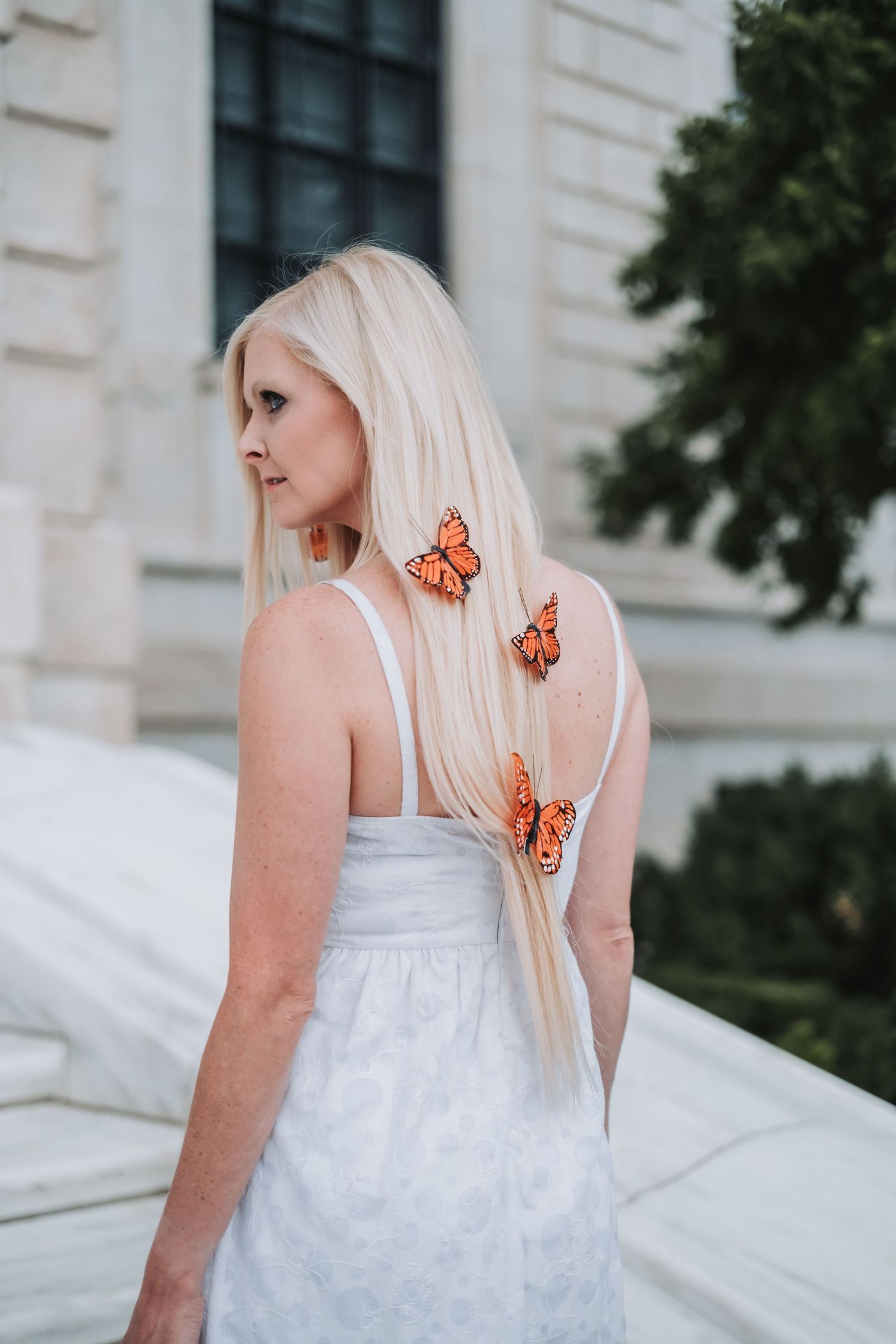 Blonde woman in white dress with orange monarch butterflies in her hair, standing outdoors near a classic building.