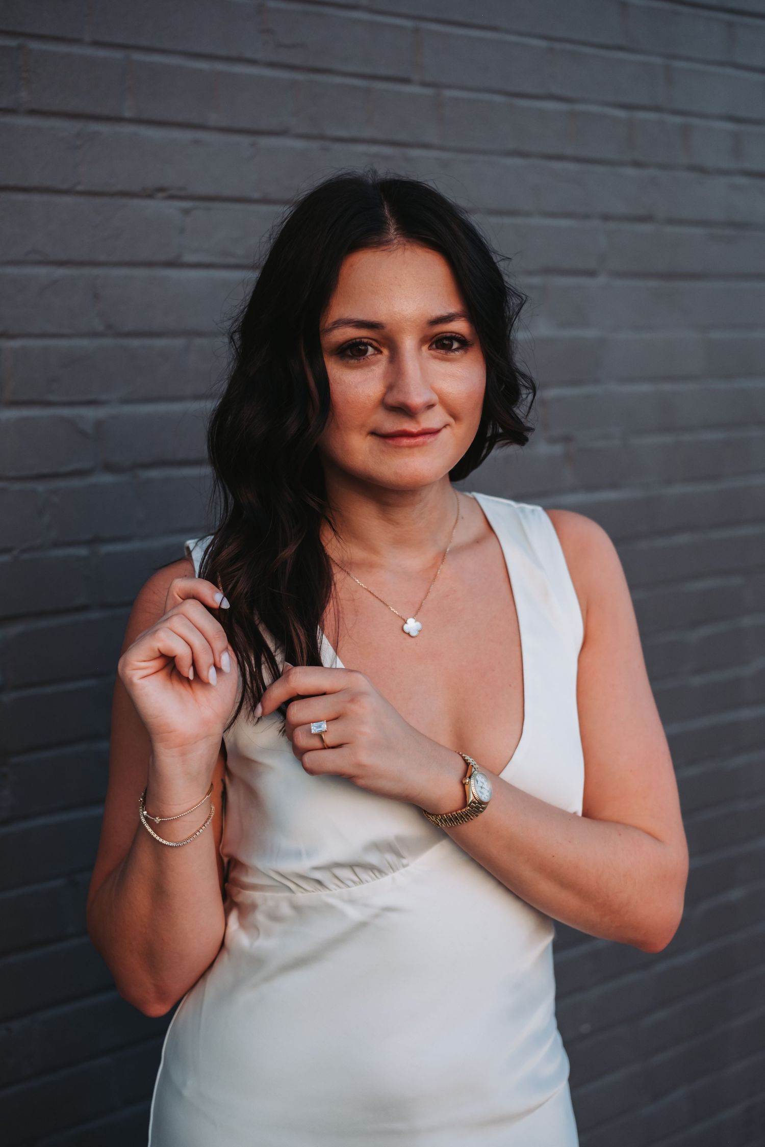 Woman in white satin dress posing against gray brick wall wearing gold jewelry and watch.