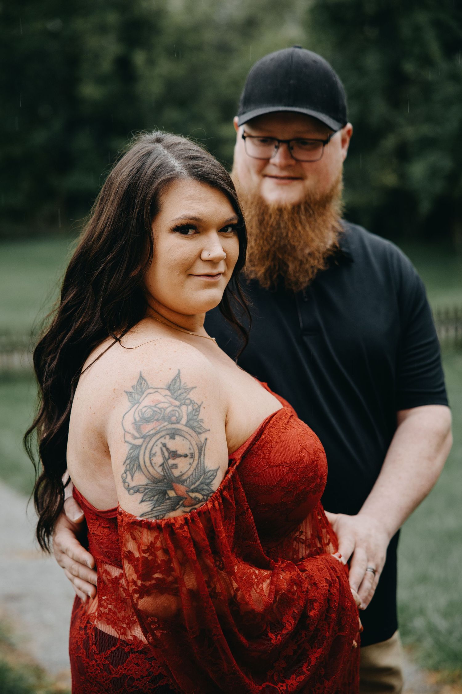 A woman in a red lace dress with a floral tattoo poses with a bearded man in a black shirt outdoors.
