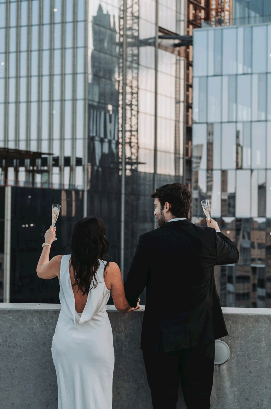 A bride and groom toast with champagne on a rooftop terrace overlooking a glass skyscraper cityscape.