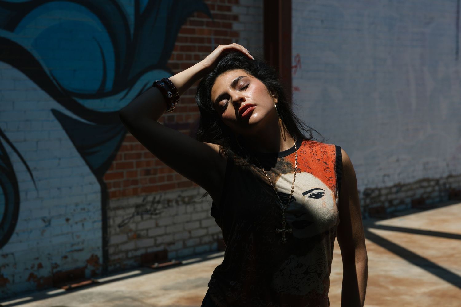 Woman in black tank top with face print poses against graffiti wall in dramatic sunlight.