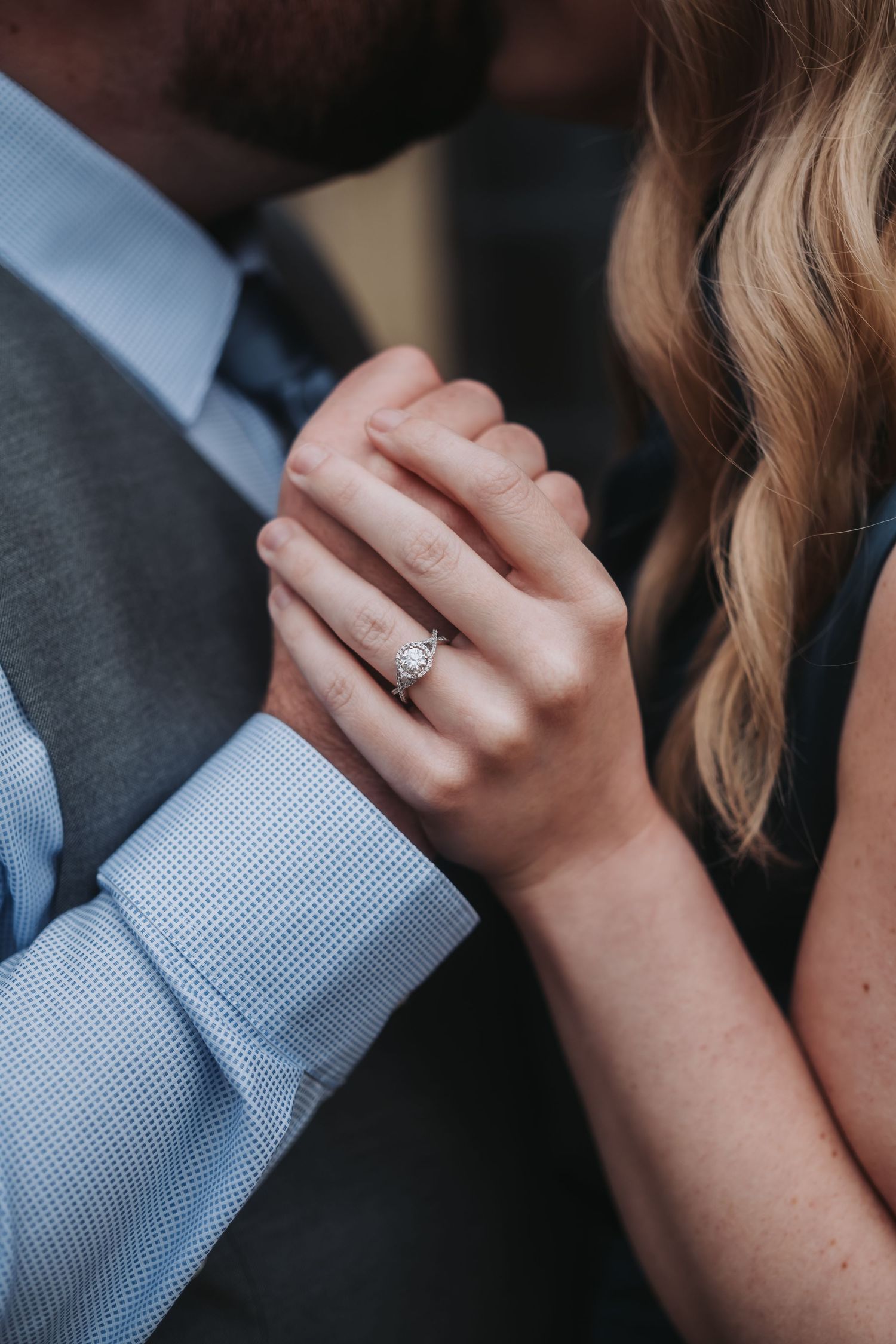 Close-up of a woman's hand wearing a diamond engagement ring while holding hands with a man in a suit.