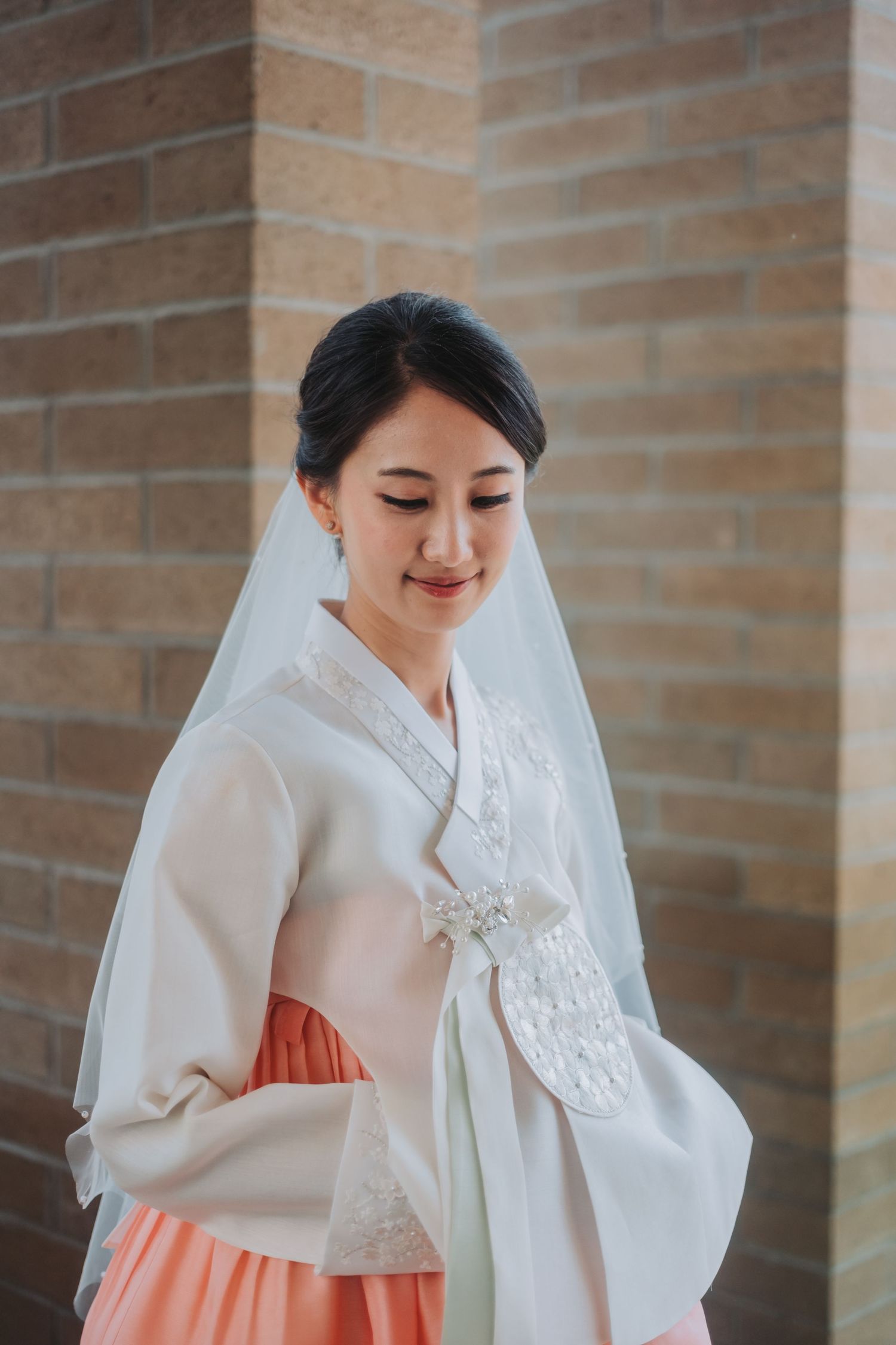 Bride in traditional white Korean hanbok poses elegantly against brick wall background.