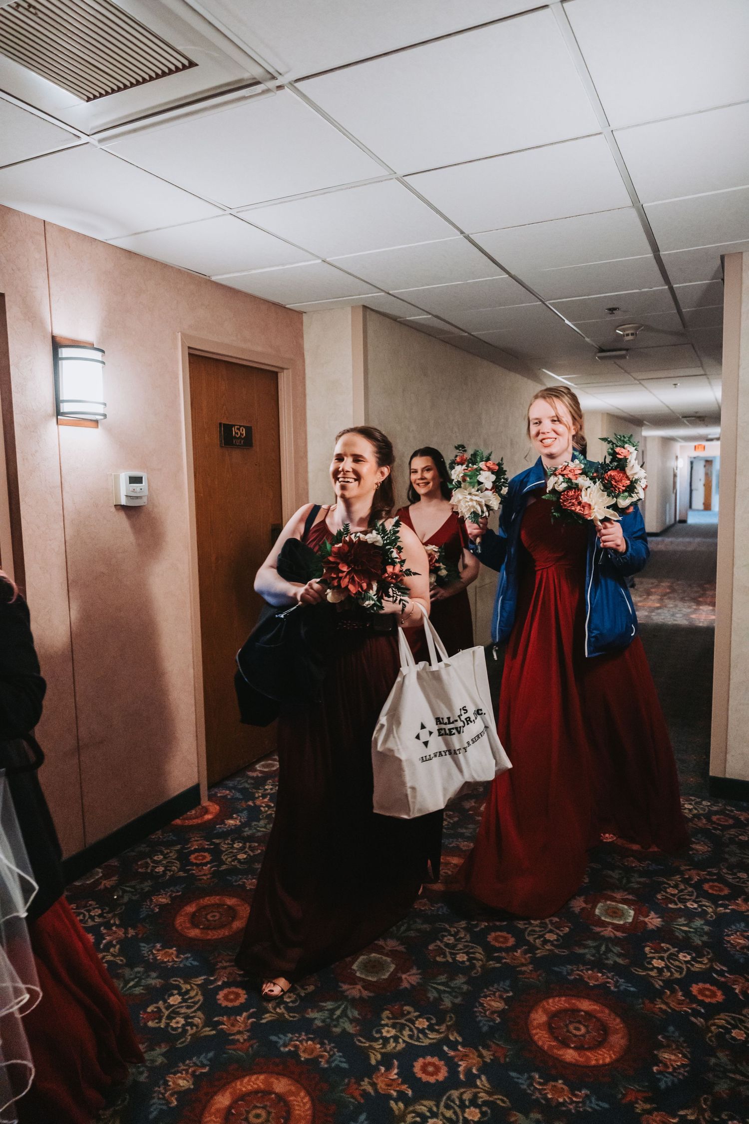Joyful wedding party members carrying bouquets and bags in a hotel hallway.