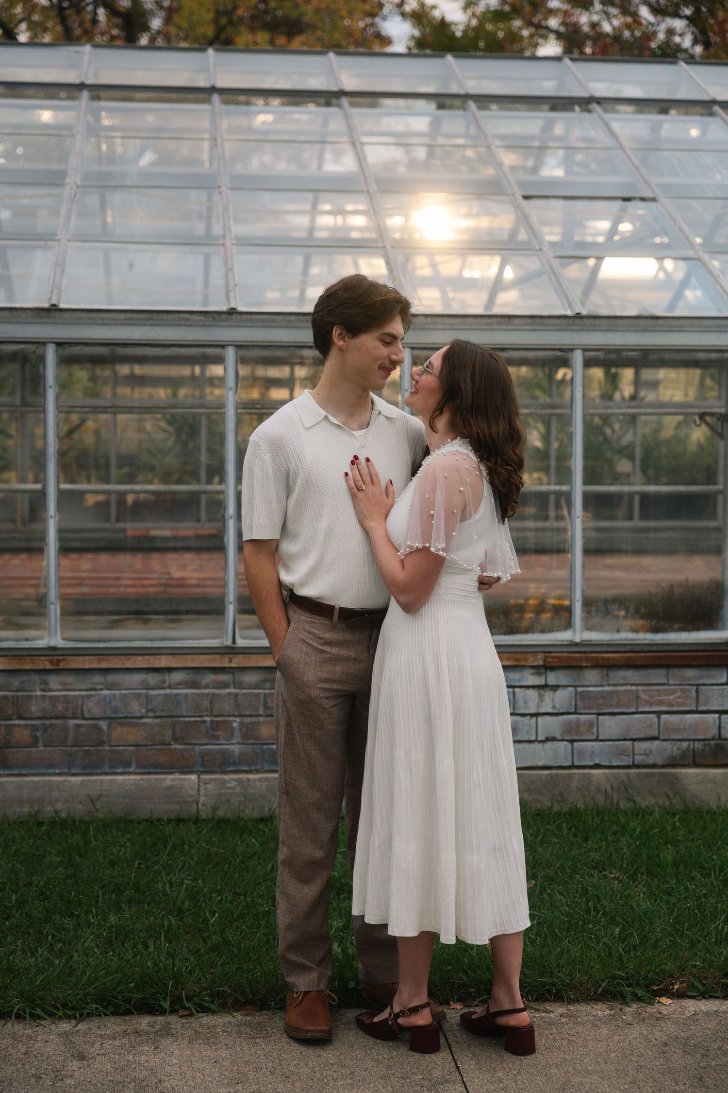 A couple shares a romantic moment in front of a glass greenhouse, woman in white midi dress, man in white polo.