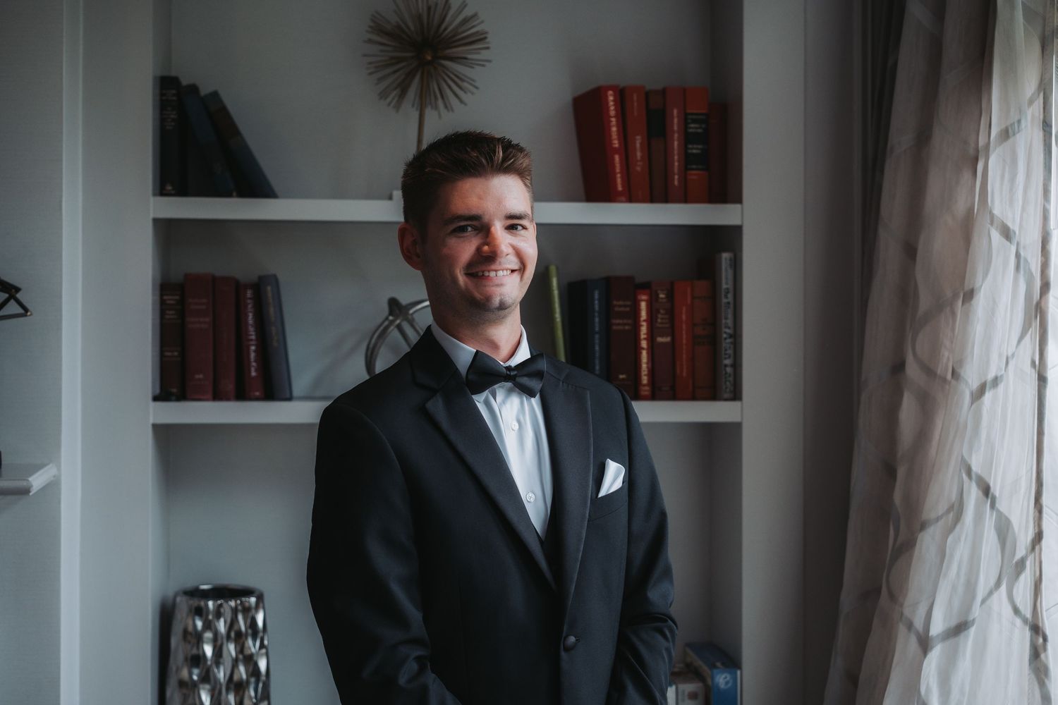 Formal dressed figure standing in moody interior room with window light.