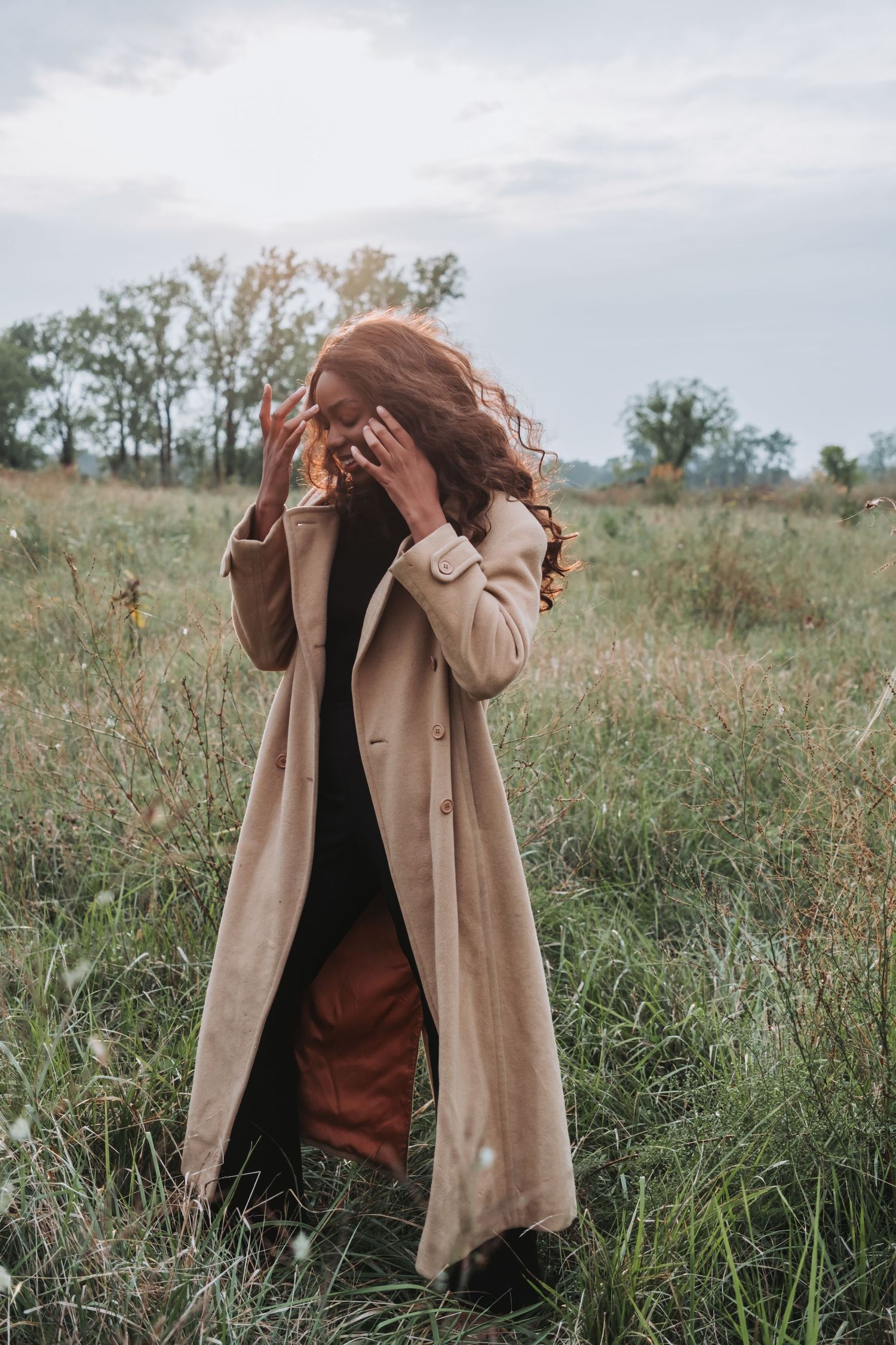 A woman with red curly hair in a beige trench coat stands in a grassy field on an overcast autumn day.