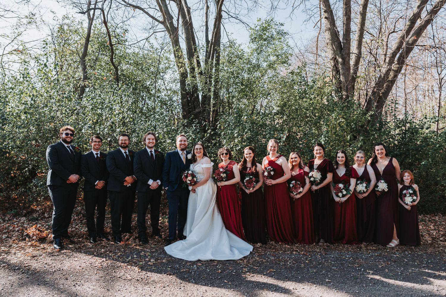 Wedding party poses outdoors, groomsmen in navy suits and bridesmaids in burgundy gowns flanking bride and groom in forest setting.