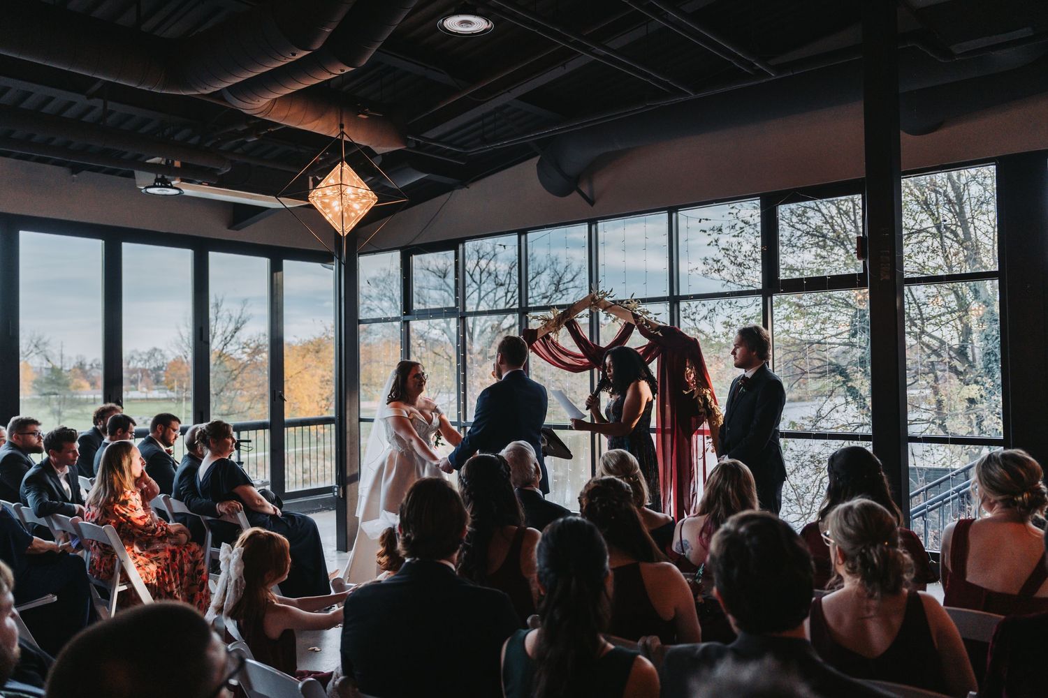 Indoor wedding ceremony with guests seated, couple at floral arch, large windows with autumn trees backdrop.