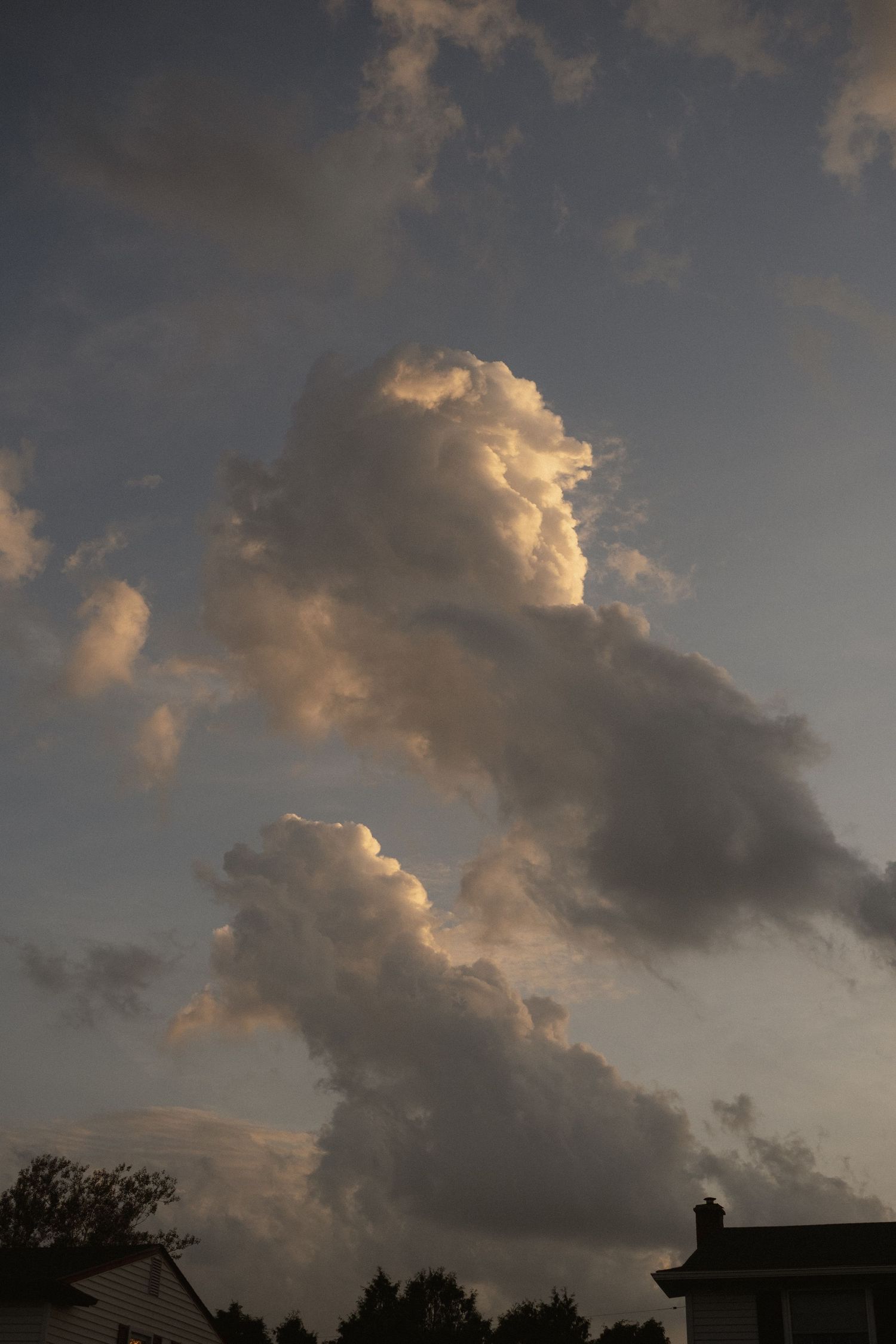 Dramatic cumulus clouds illuminated by golden sunset light tower over suburban rooftops against a moody gray sky.