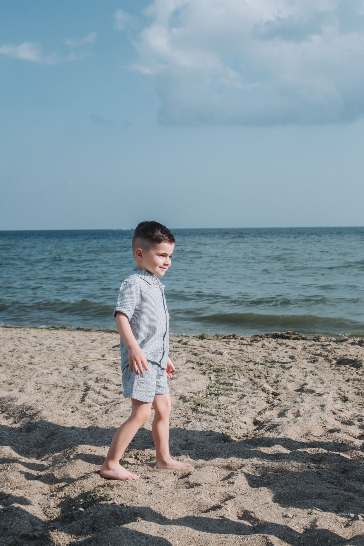A young boy in a gray outfit walks barefoot on a sandy beach with calm blue ocean waters in the background.