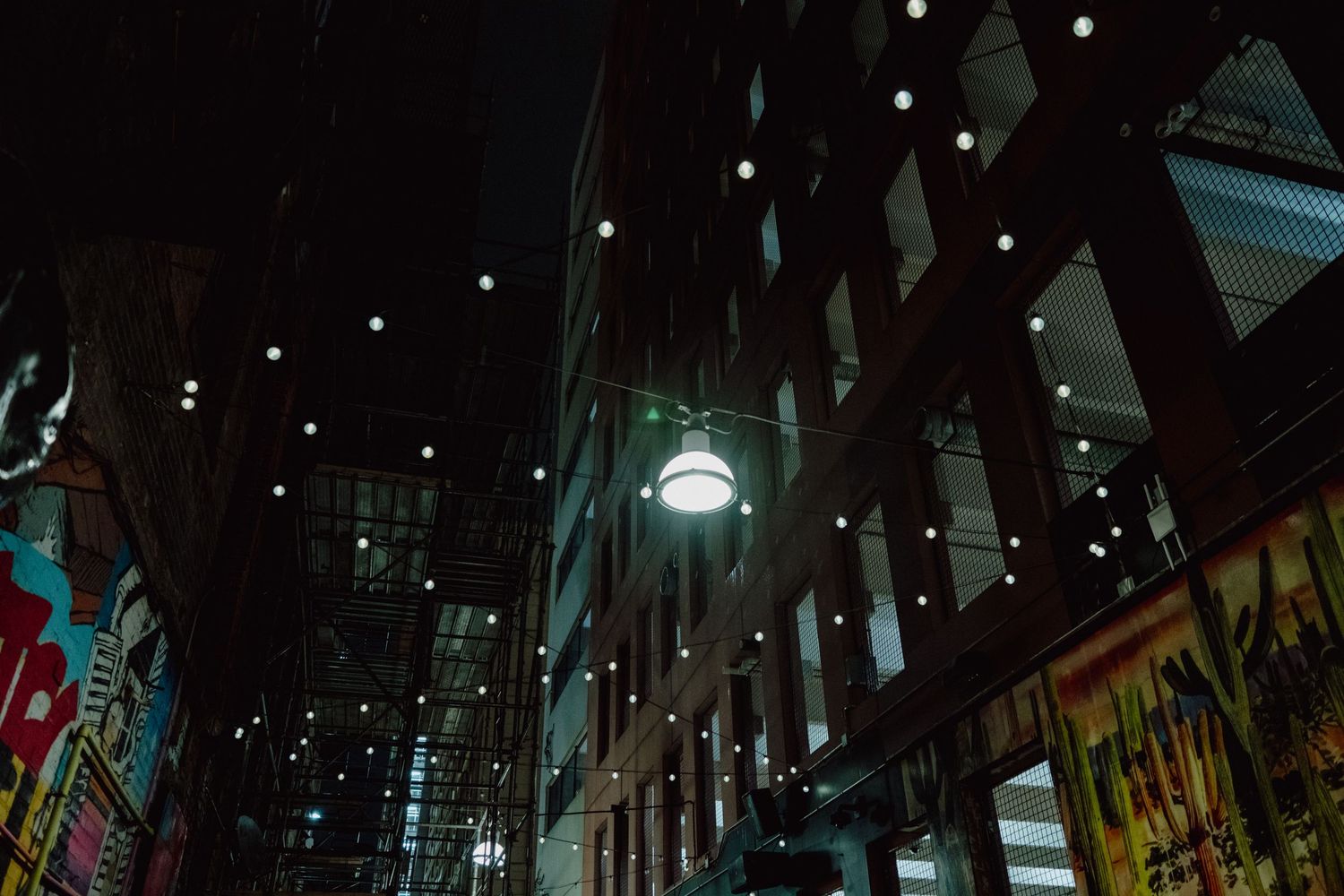 Dark urban alley lit by string lights and a glowing globe lamp, with colorful graffiti murals on brick walls at night.