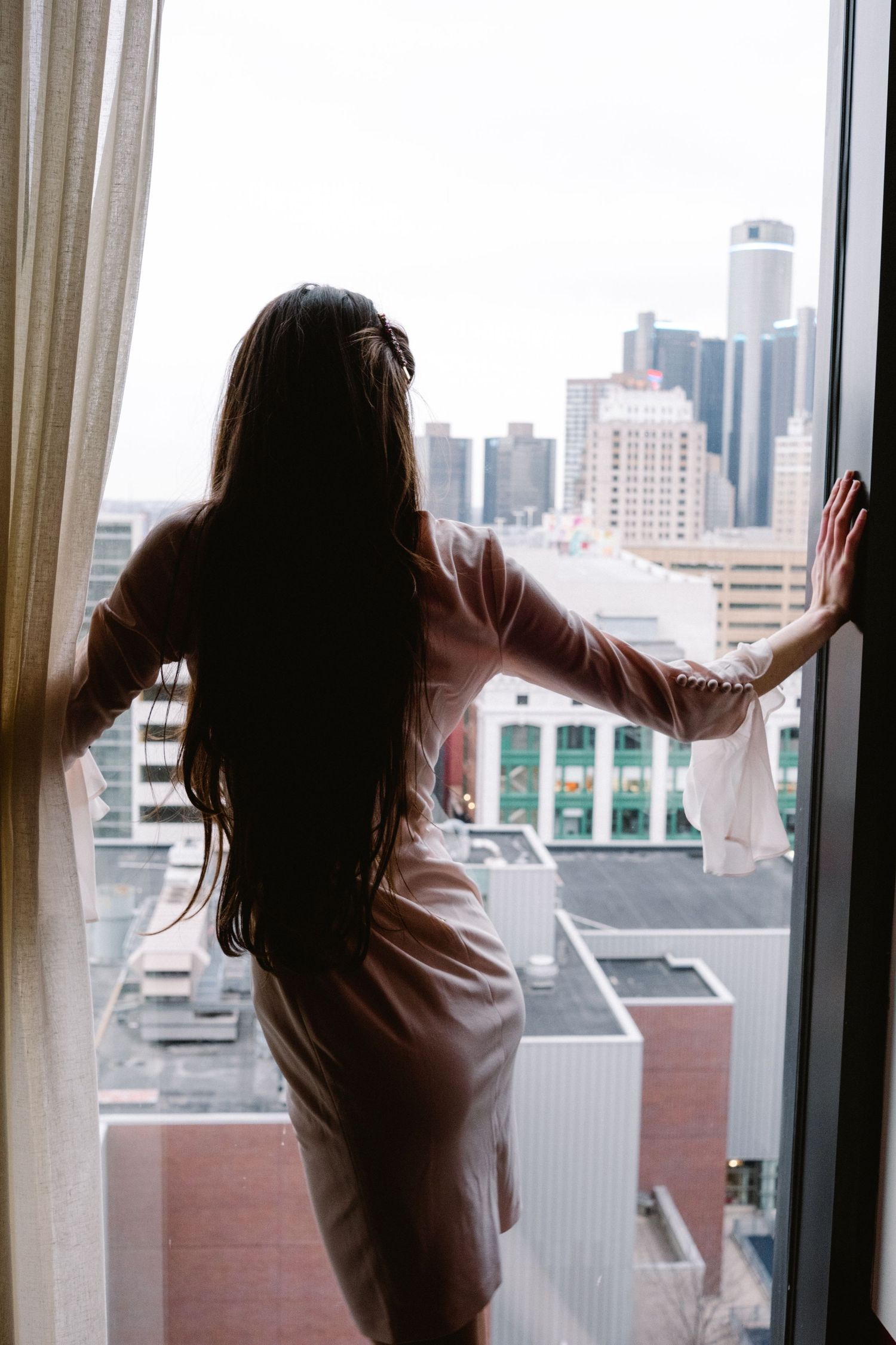 Woman in silk dress gazes at city skyline through hotel window, long dark hair flowing, urban buildings visible outside.