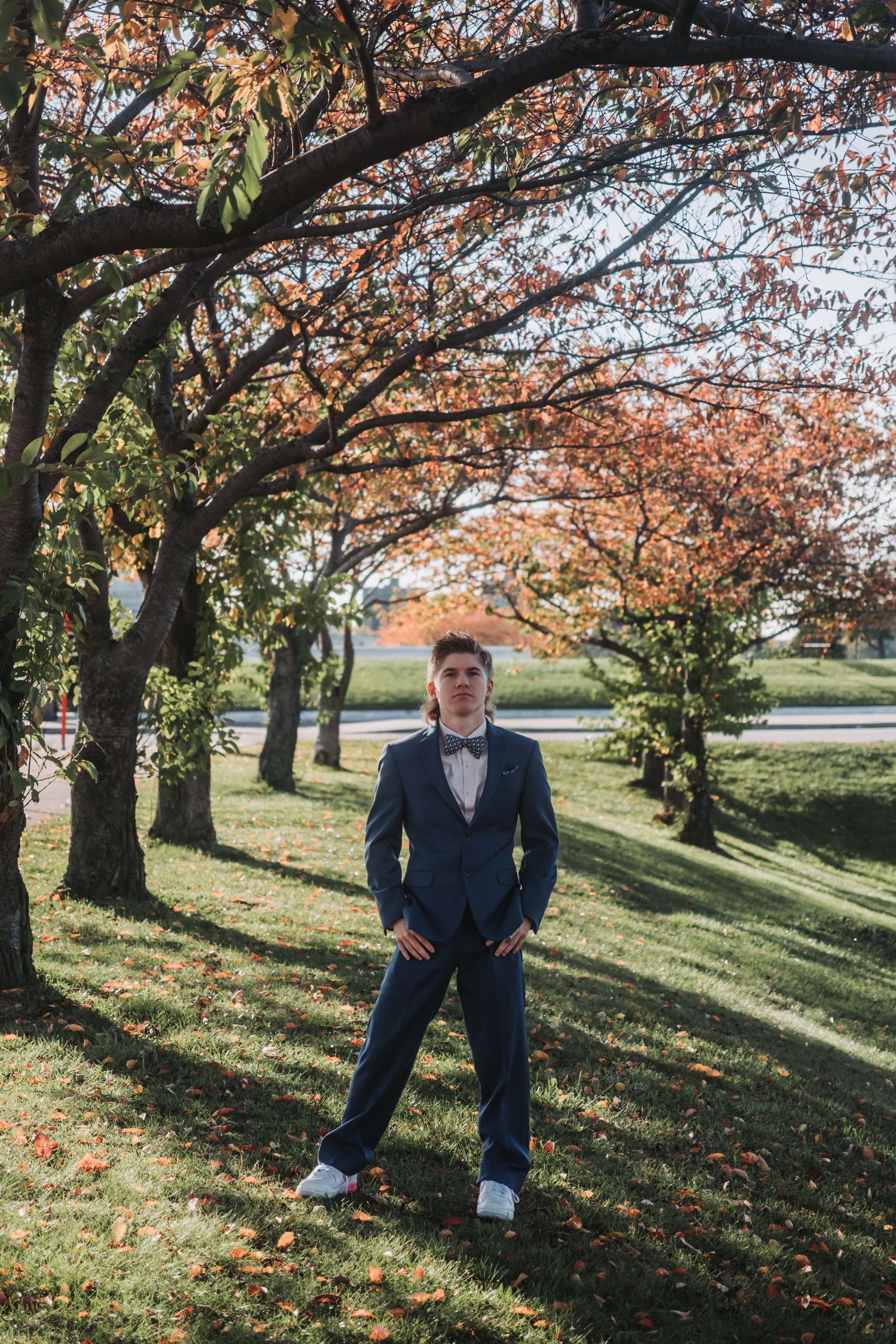Young man in navy suit and bow tie poses confidently under autumn trees in a park setting.