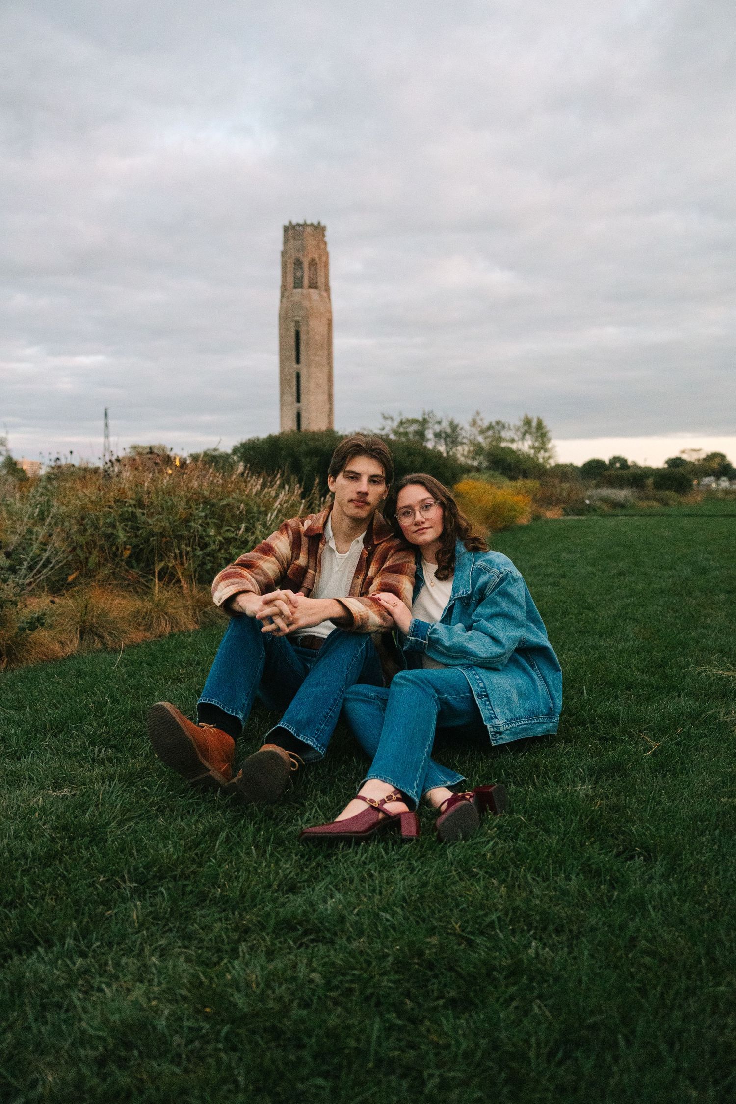 Couple sitting together on green grass with a historic tower in the background under a cloudy sky.