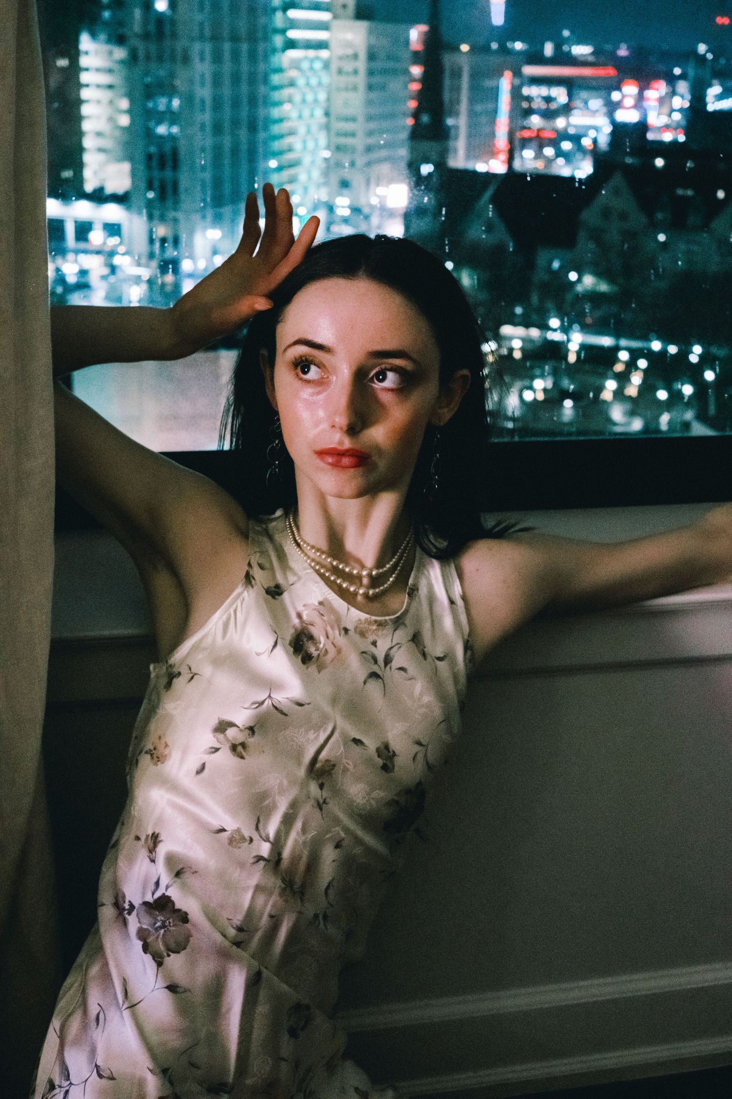 Young woman in floral dress and pearl necklace poses by a window with city lights at night.