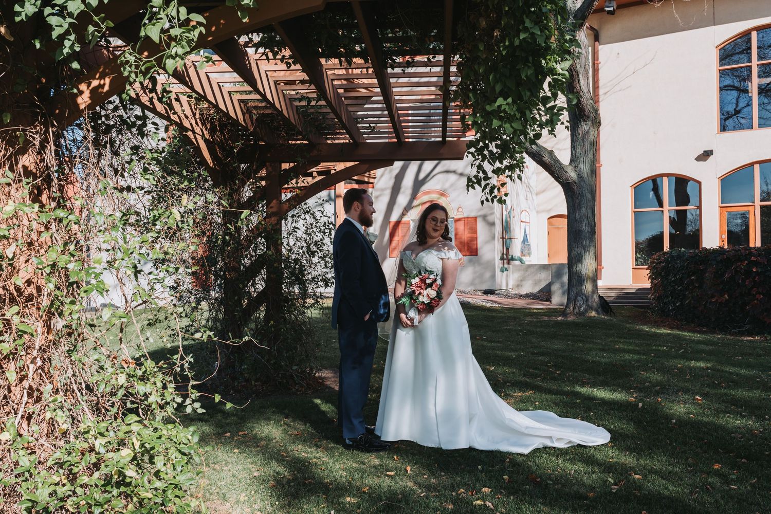 A wedding couple stands beneath a pergola in a garden setting on a sunny afternoon.