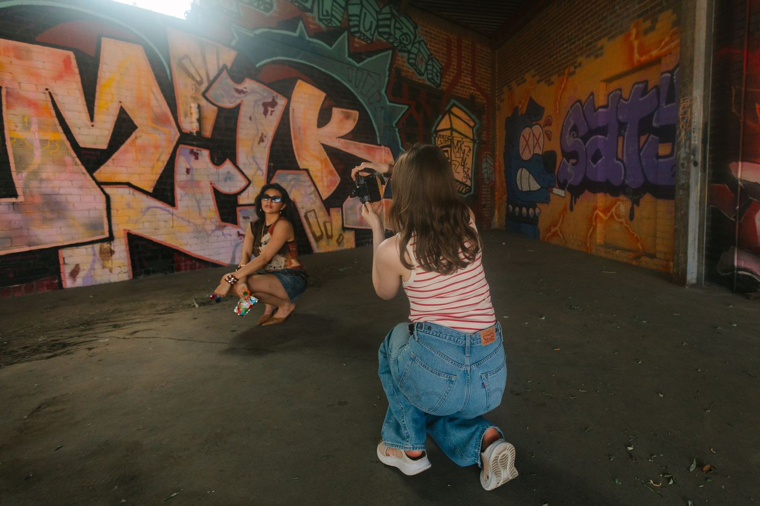 Two girls photographing each other in a colorful graffiti-covered urban space, one posing while the other kneels with a camera.