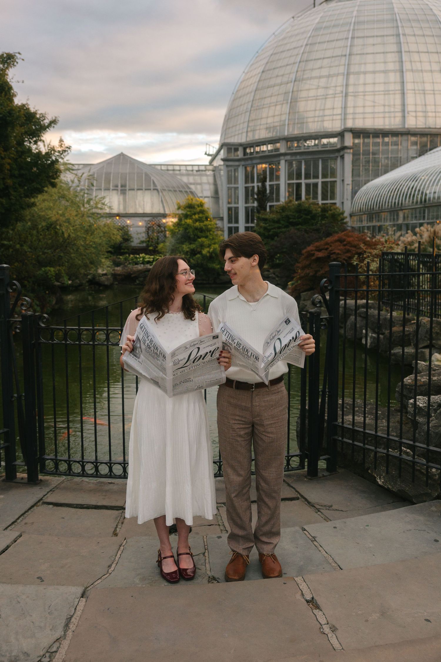 A couple poses together in front of a glass conservatory dome during an engagement photo session at sunset.