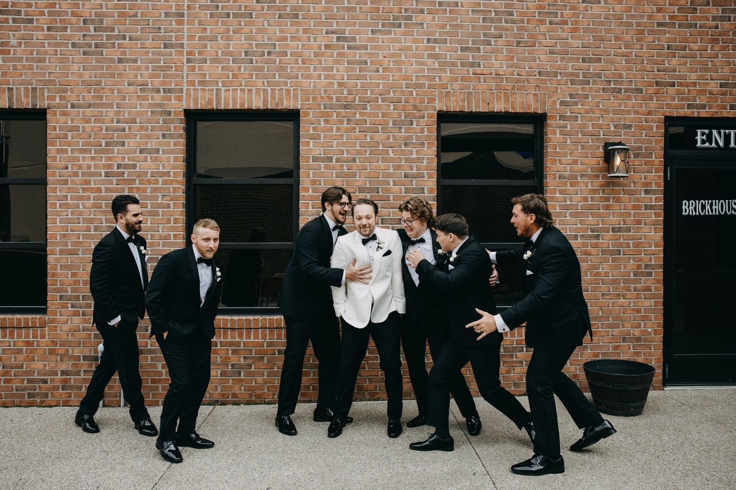 Groom in white tuxedo laughing with groomsmen in black tuxedos outside brick building at wedding.