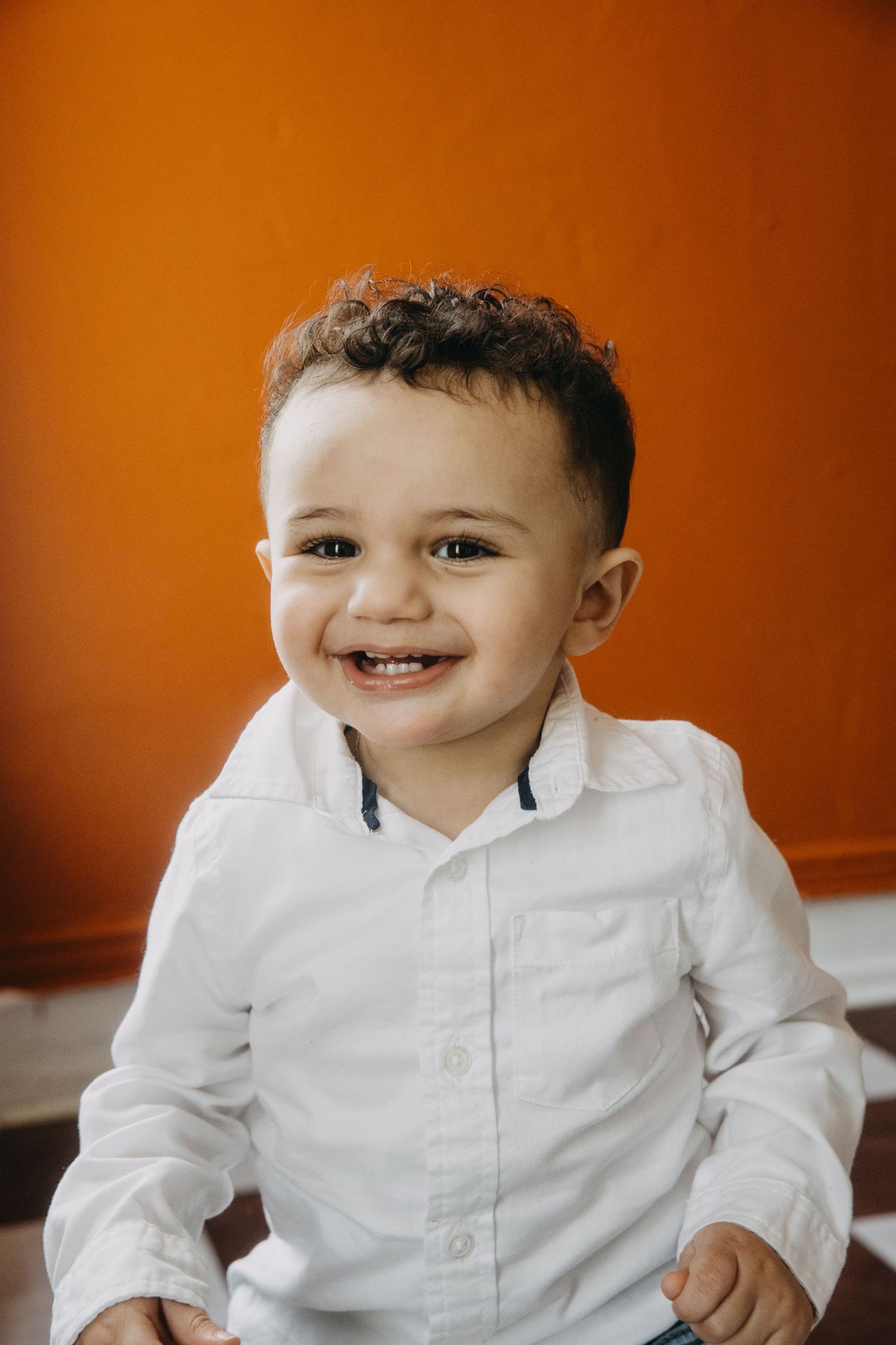 Smiling toddler boy in white button-up shirt against bright orange wall.