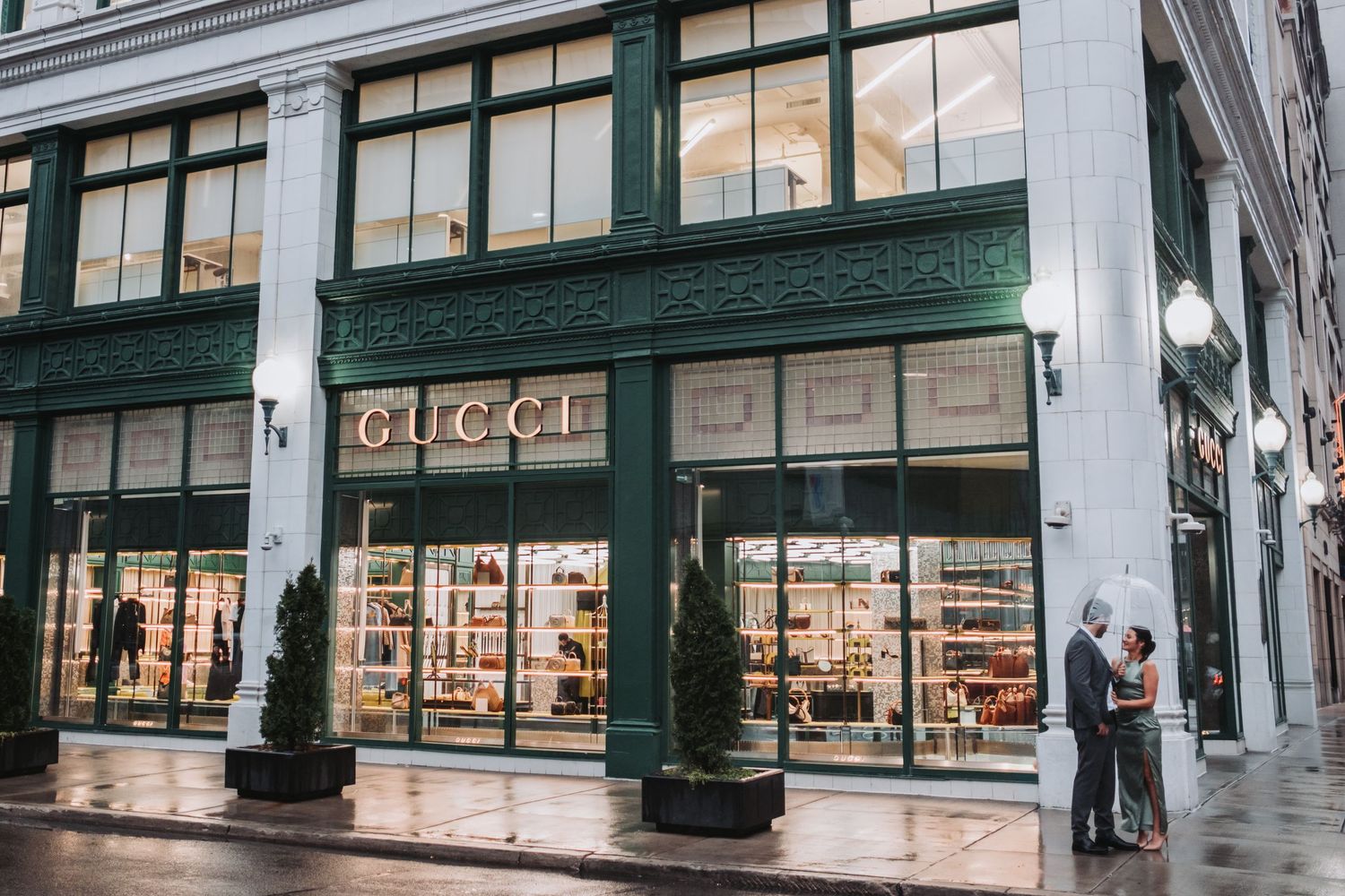 Gucci luxury retail store exterior with green facade, large display windows, and shoppers on a rainy city street.