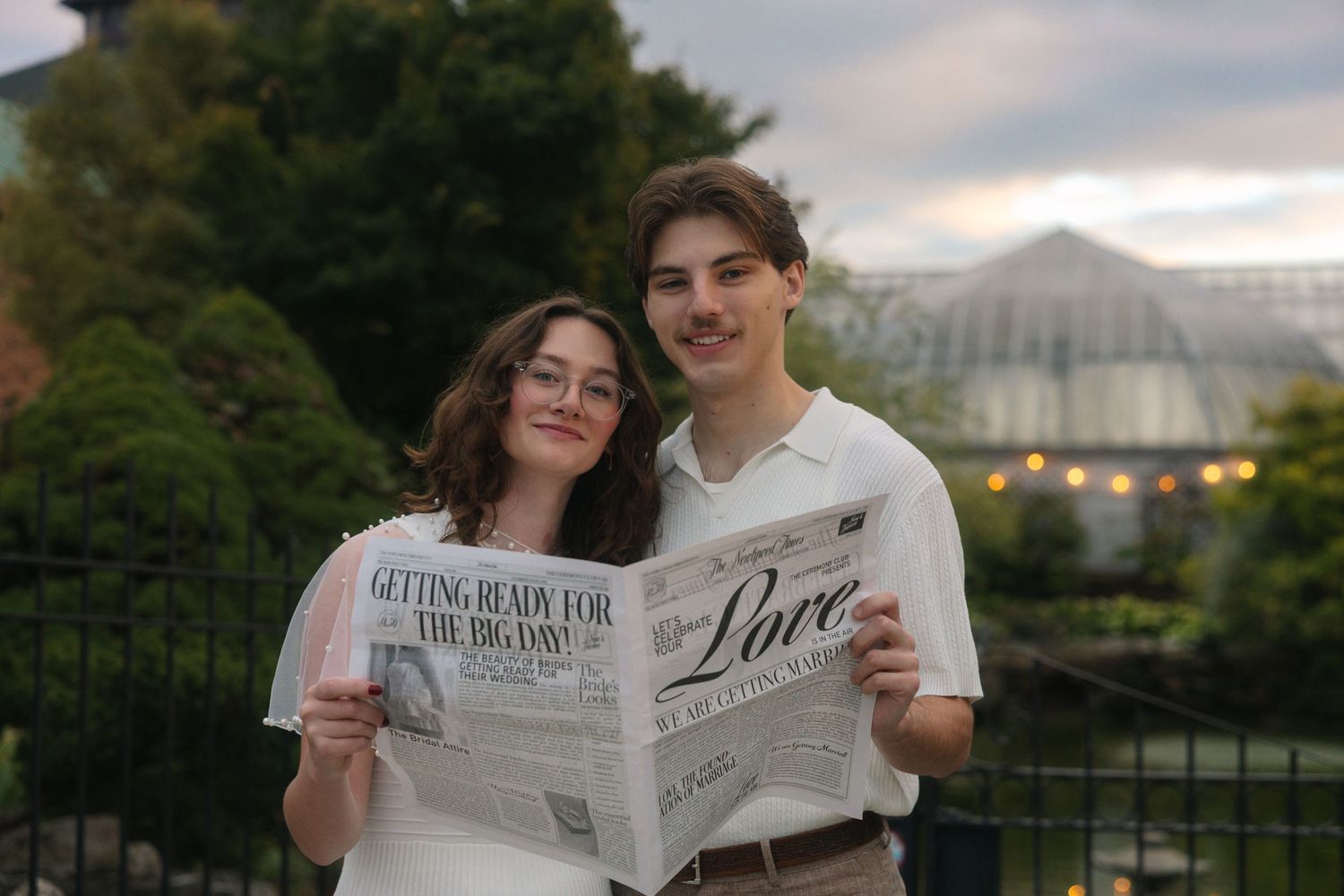 Couple smiling and holding a Love newspaper outdoors near a greenhouse at dusk.