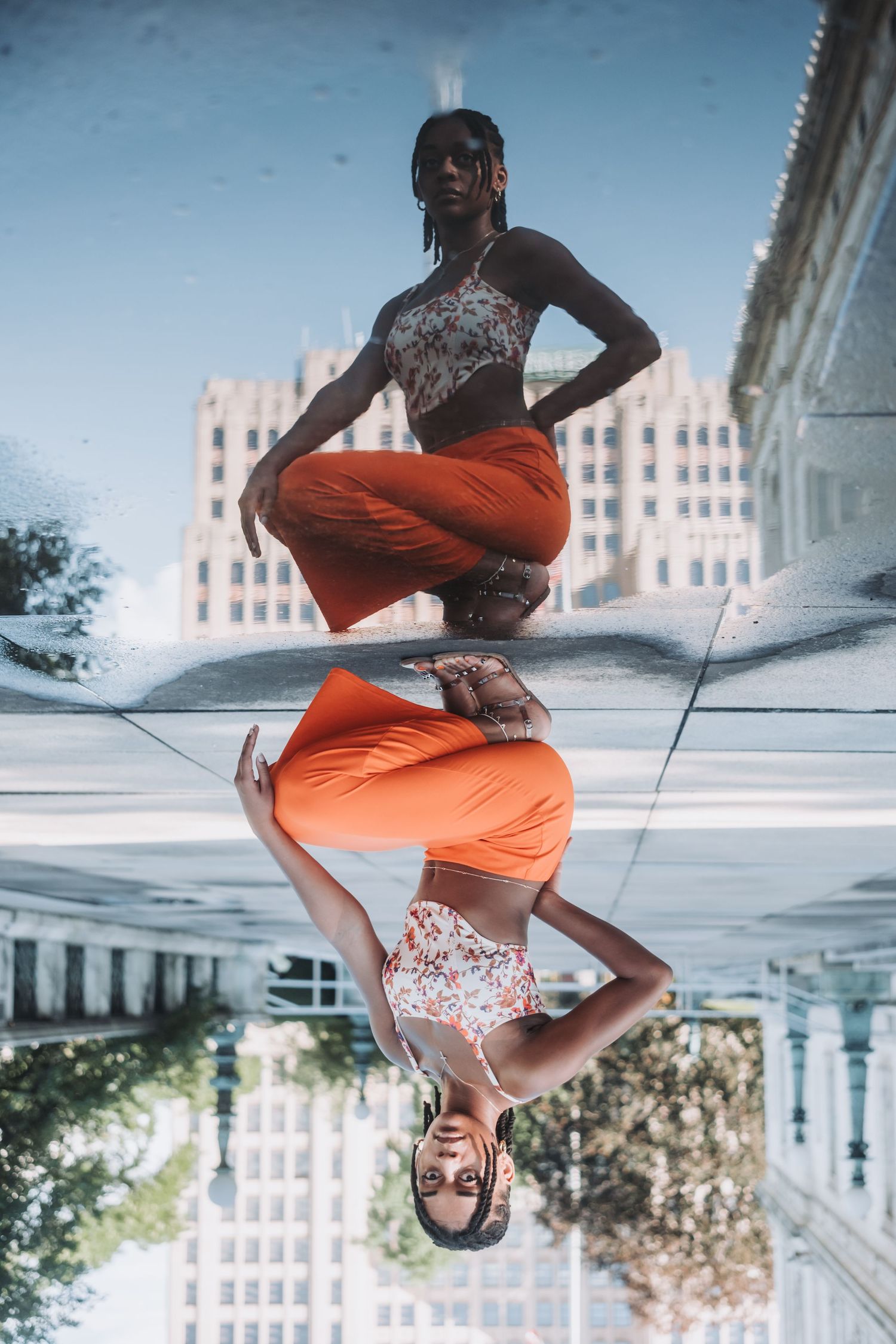 Woman in orange pants and floral top posing outdoors, reflected in a mirrored surface with city buildings behind her.