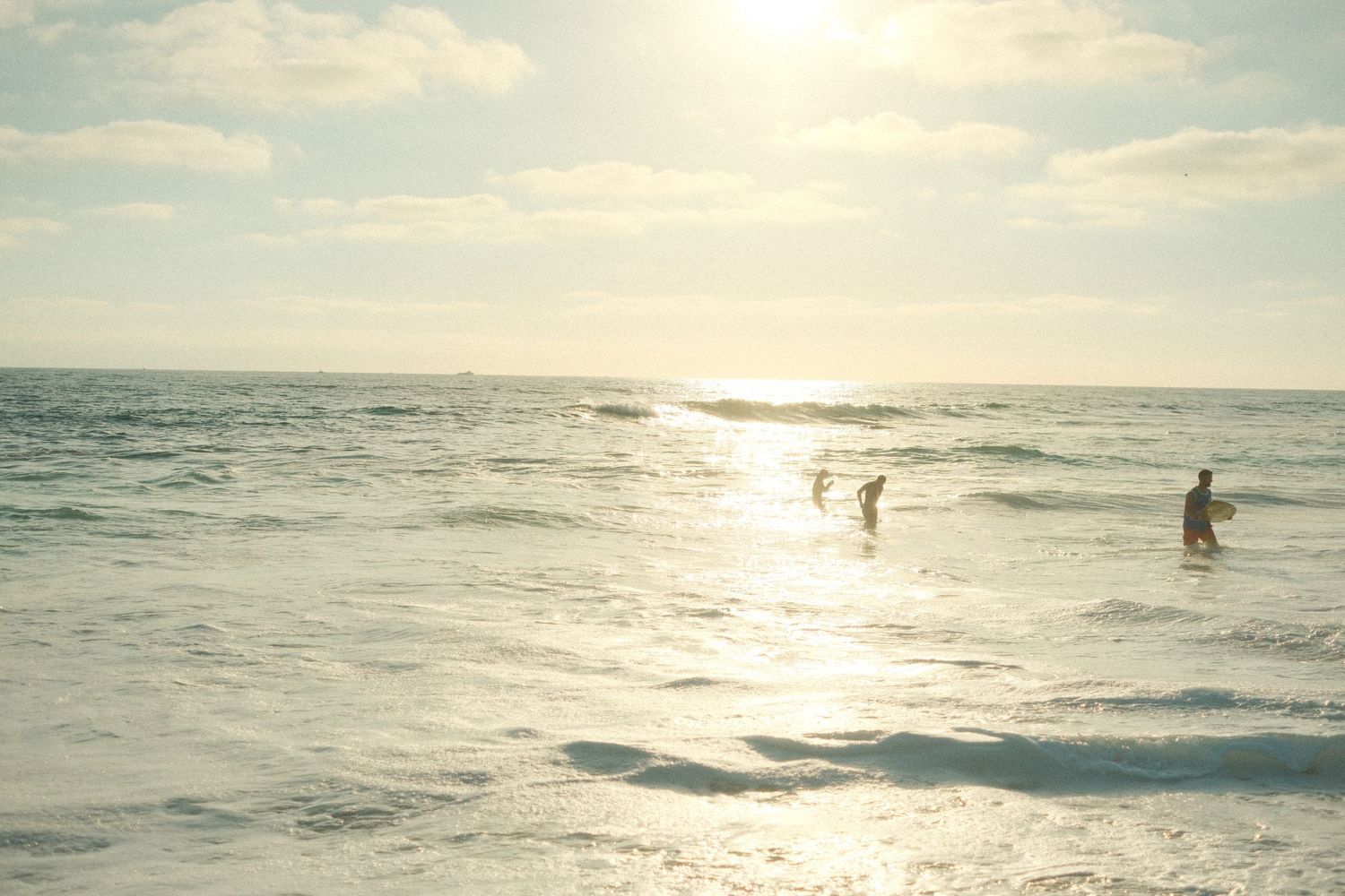 People wading in sunlit ocean waves on a bright beach day with glowing horizon and scattered clouds.