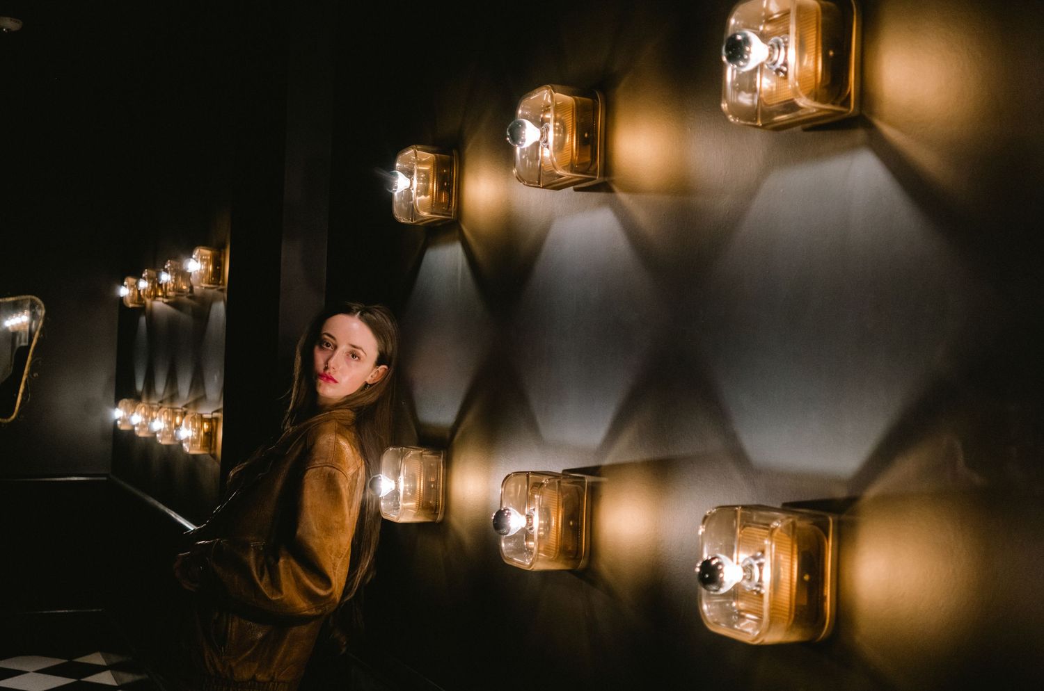 Woman standing beside glowing geometric wall sconces in a dimly lit hexagonal patterned corridor.