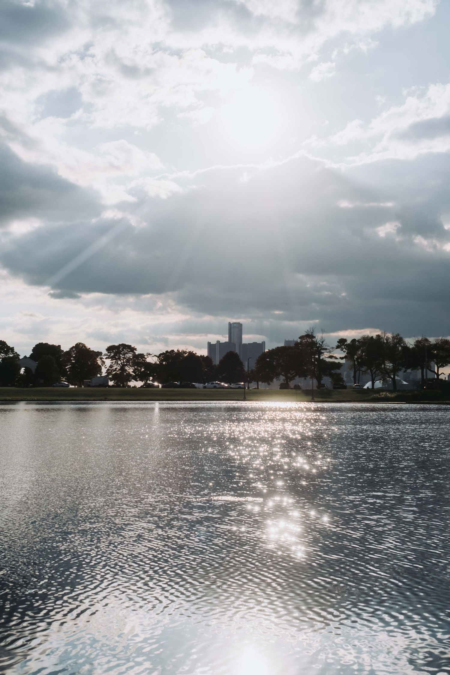 Sunlight reflecting on calm lake water with city skyline silhouette and dramatic cloudy sky in background.