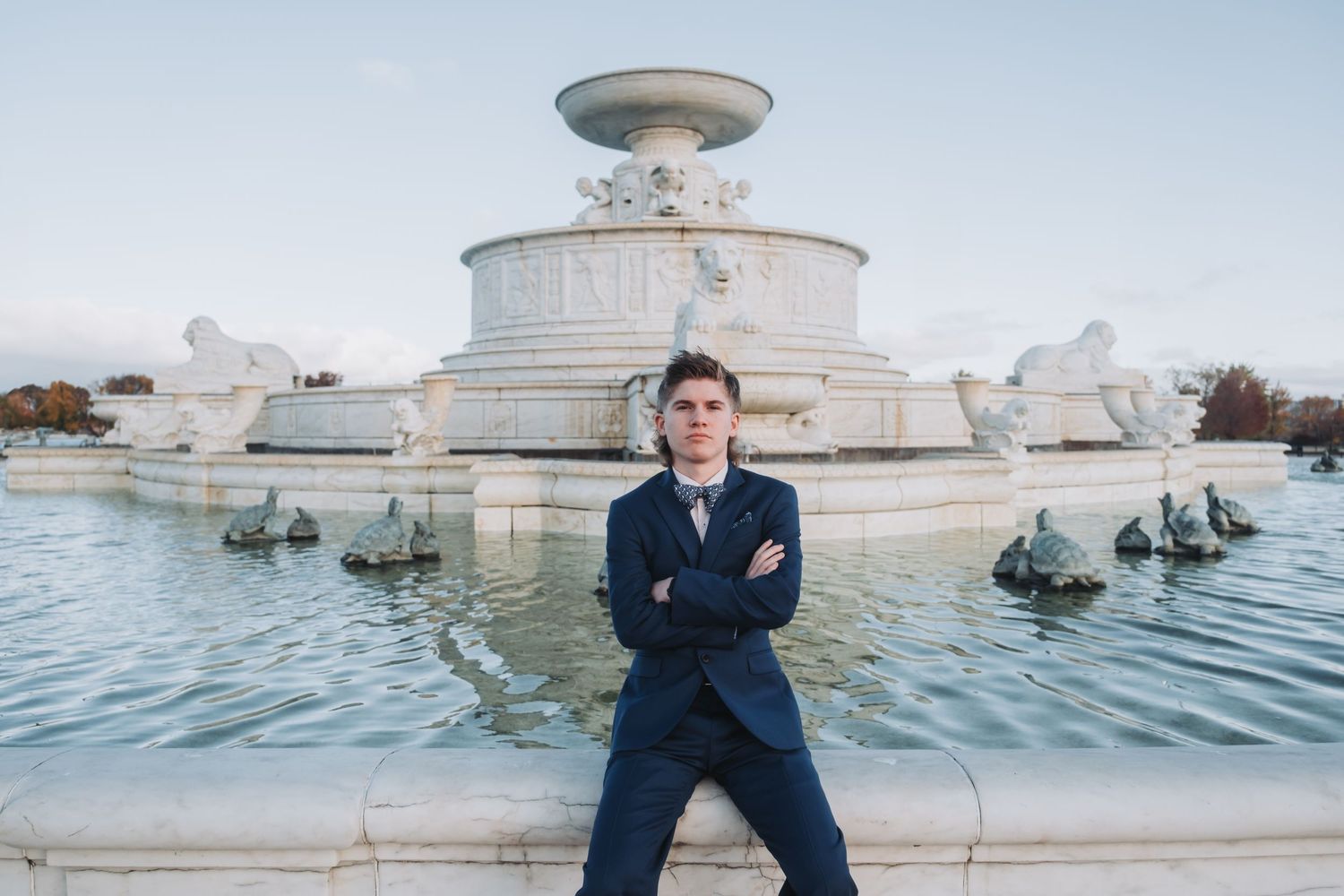 A person in navy blue stands near a fountain with classical architecture in the background.