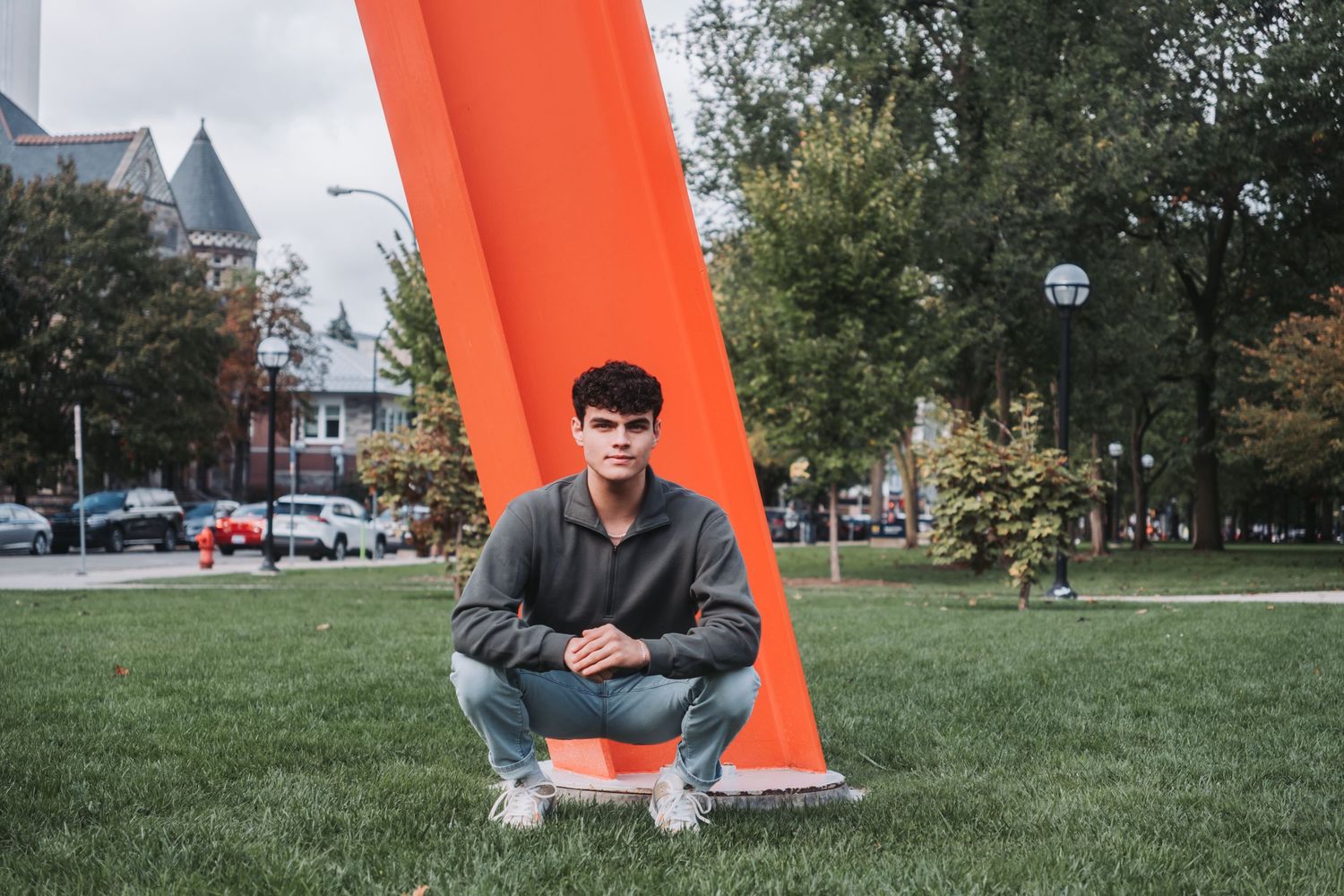 Young man crouching on grass beside a large orange sculpture in an urban park setting.