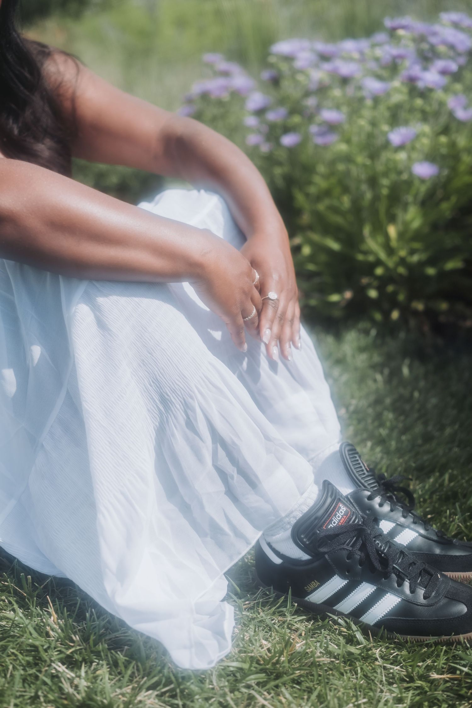 A woman in a white maxi skirt and black Adidas sneakers sits on grass near purple wildflowers.