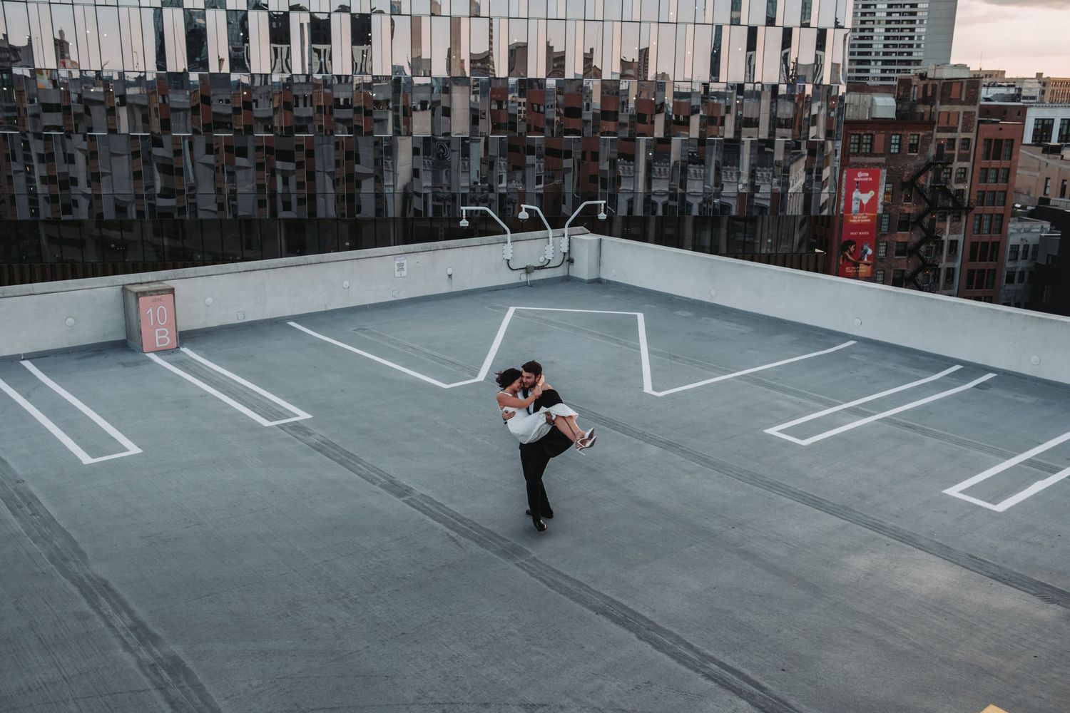 Couple embracing romantically on a rooftop parking lot with city buildings in the background.