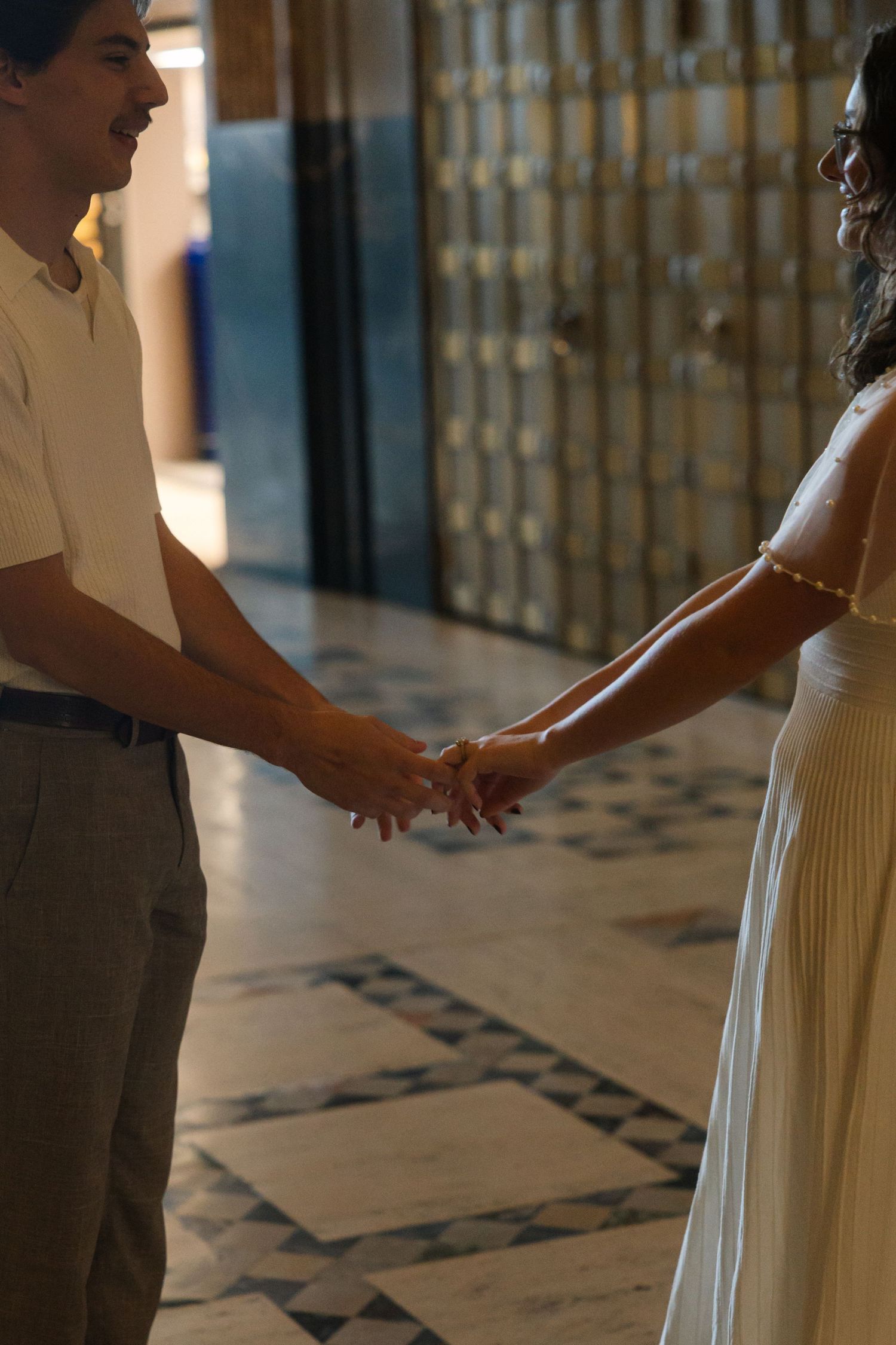 Couple holding hands in elegant hallway with marble floors and lockers, wearing formal attire.