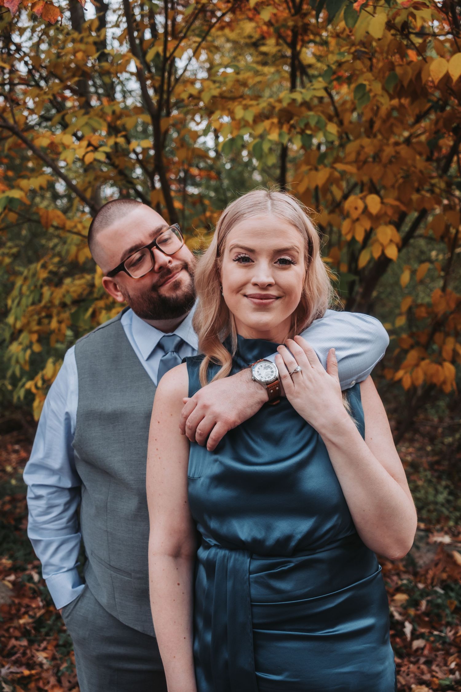 Couple poses in autumn park, man in gray vest and tie embraces woman in teal dress, engagement ring visible.