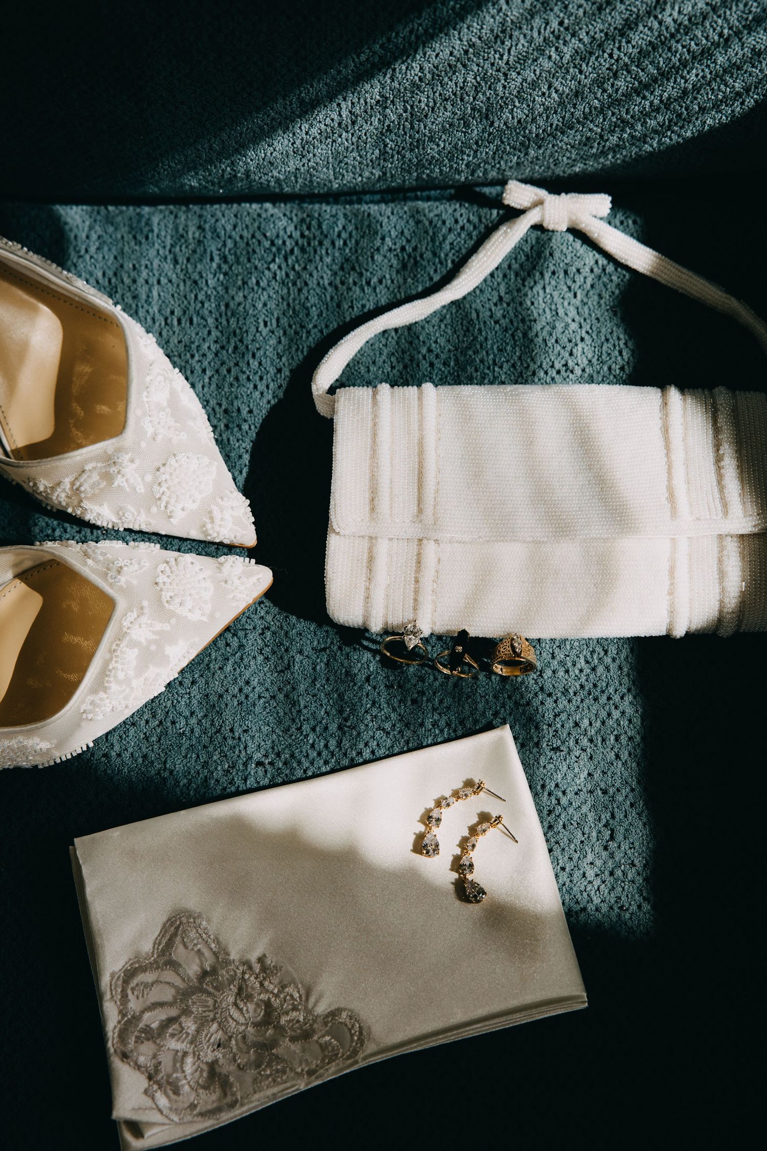 Bridal accessories flat lay featuring lace heels, white clutch purse, gold earrings, and embroidered handkerchief on teal fabric.