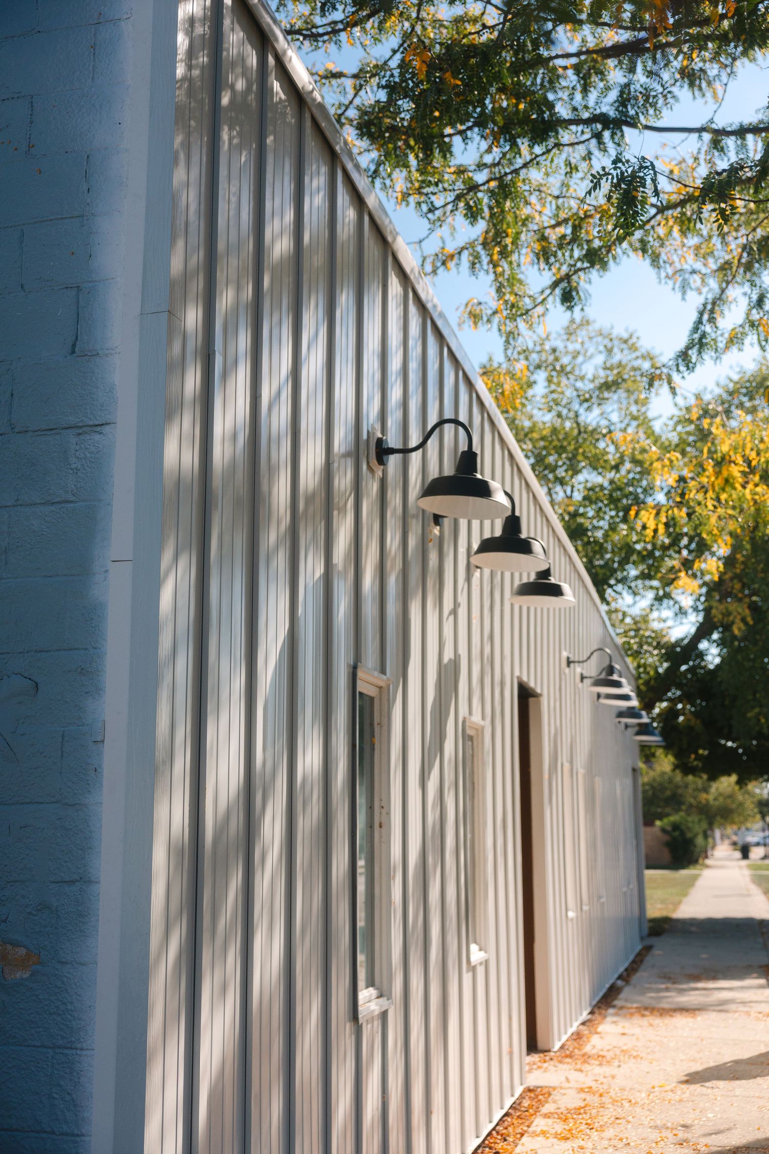 White wooden storage units with black light fixtures line an outdoor walkway beneath autumn trees.