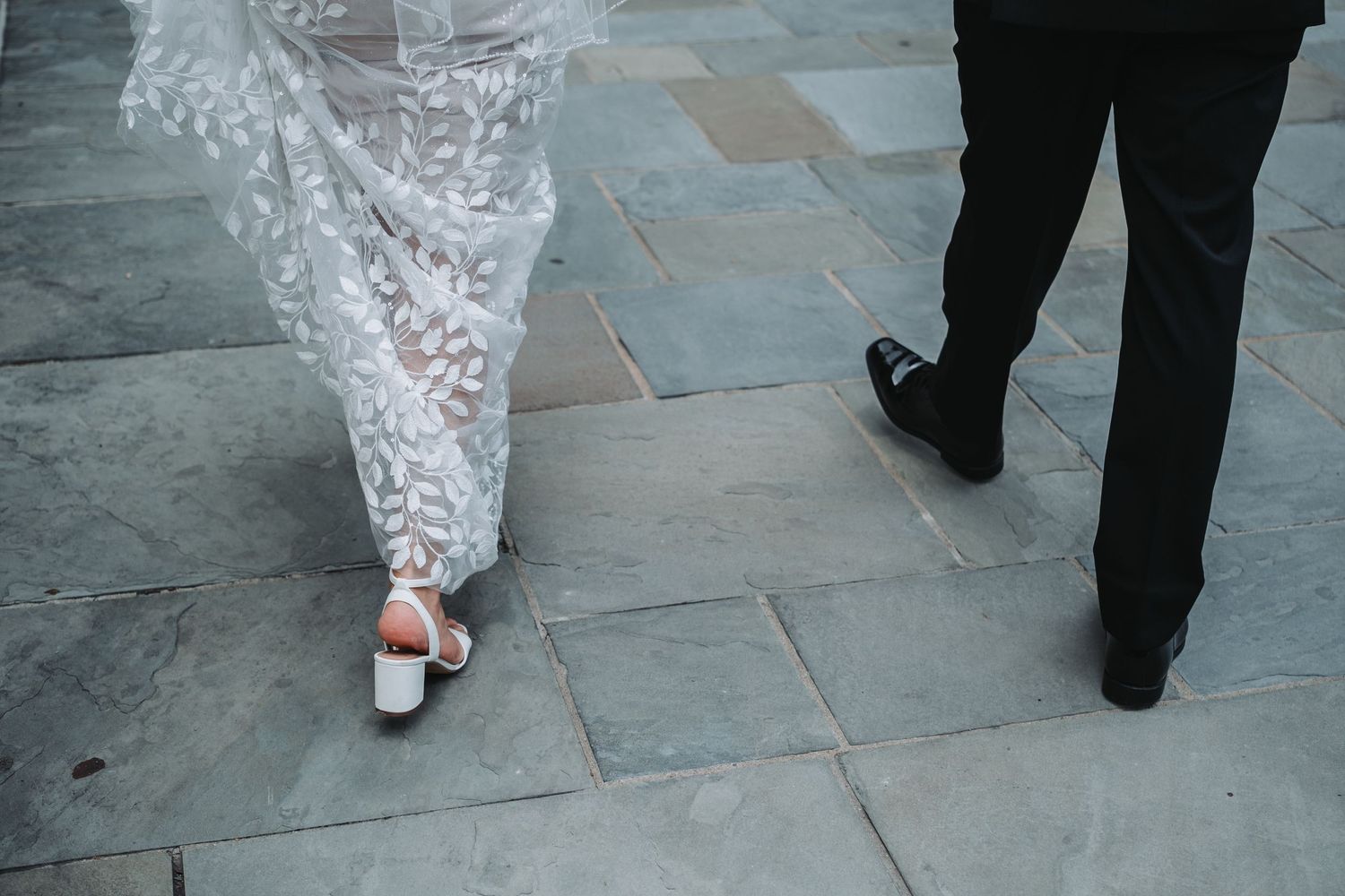 Close-up of formal wedding attire and shoes walking on stone pavement.