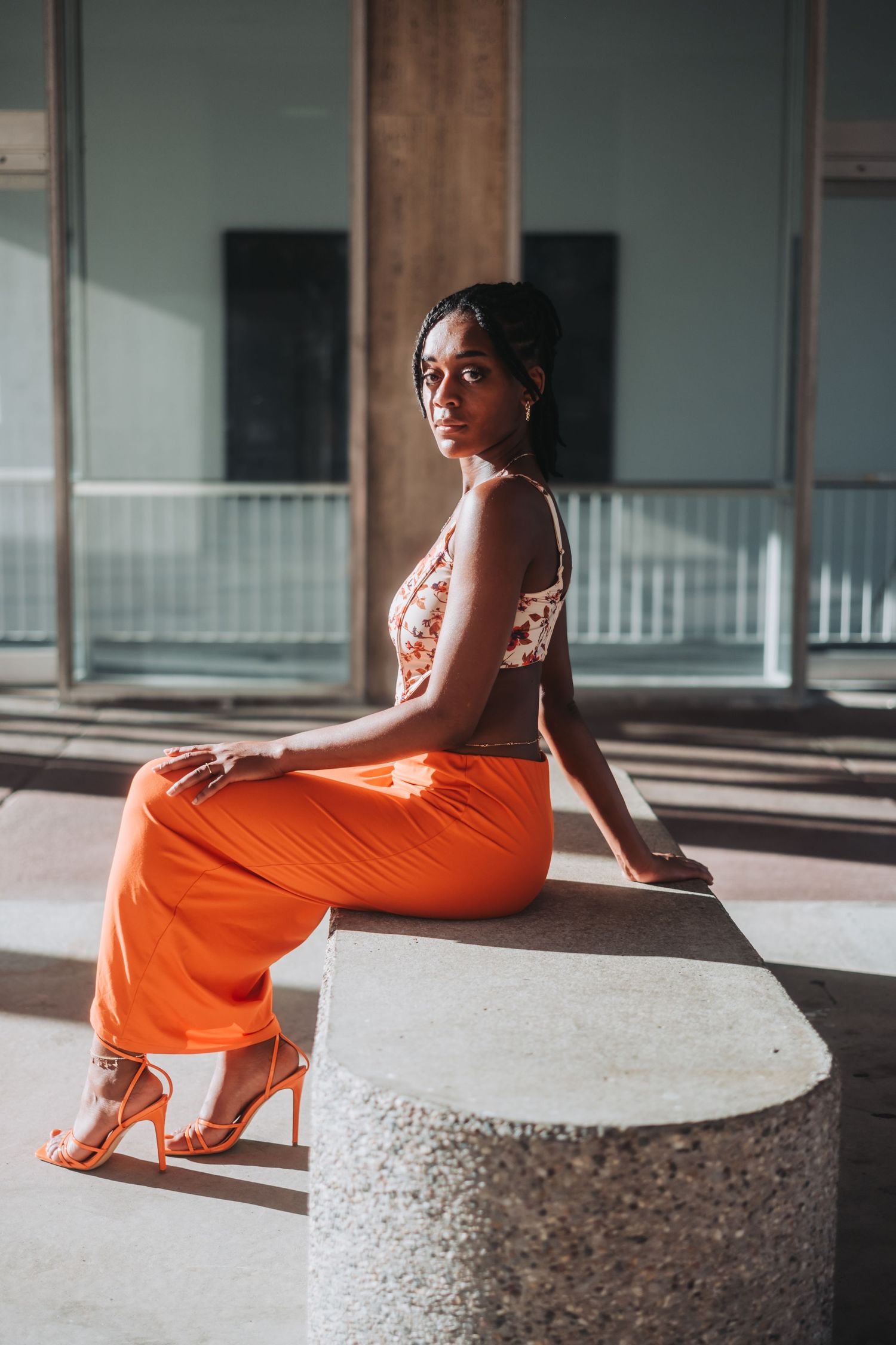 Stylish woman in orange pants sits on concrete bench in modern architectural setting with dramatic lighting.