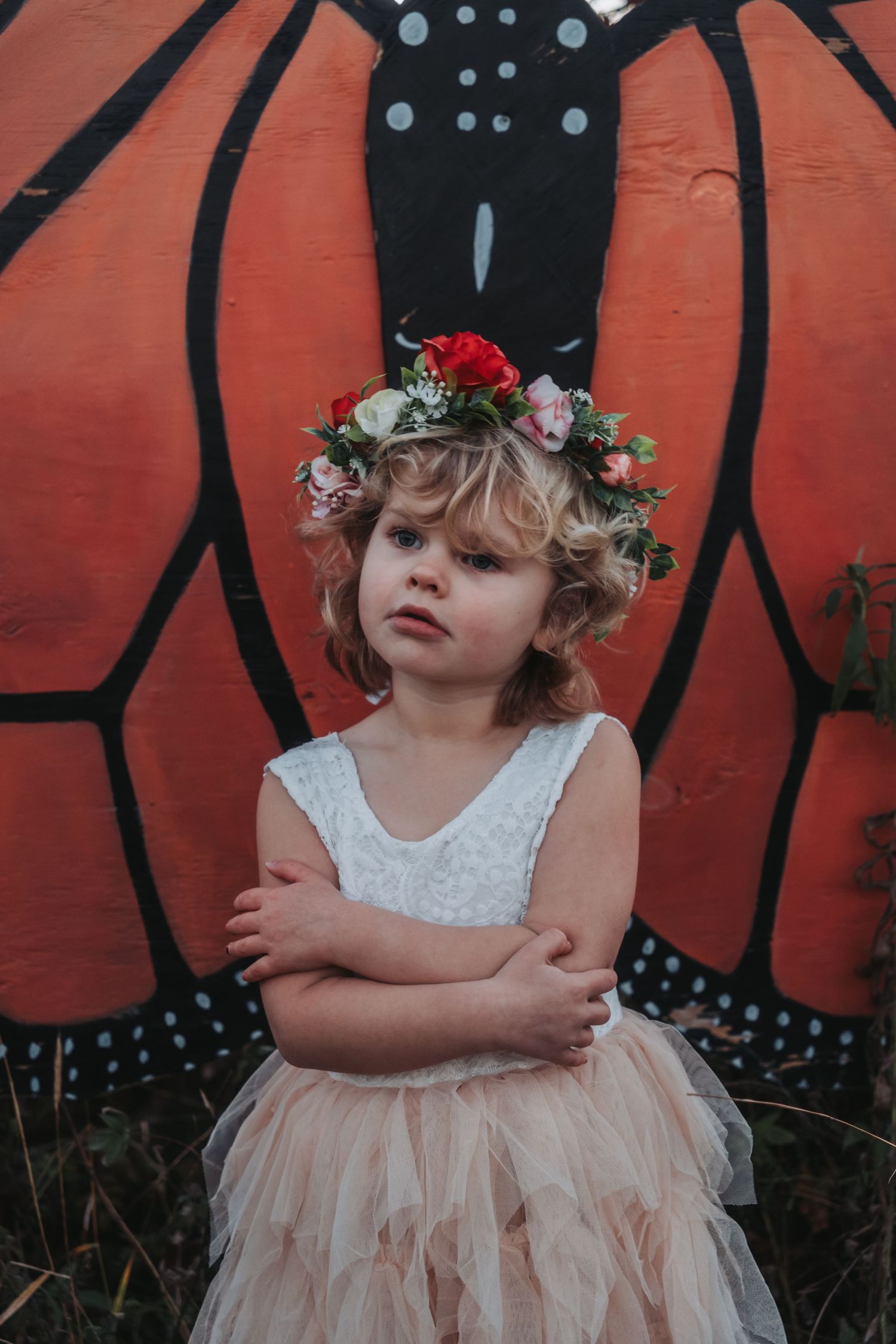 A young child in a pink tulle dress and floral crown poses against a red wall with black designs.