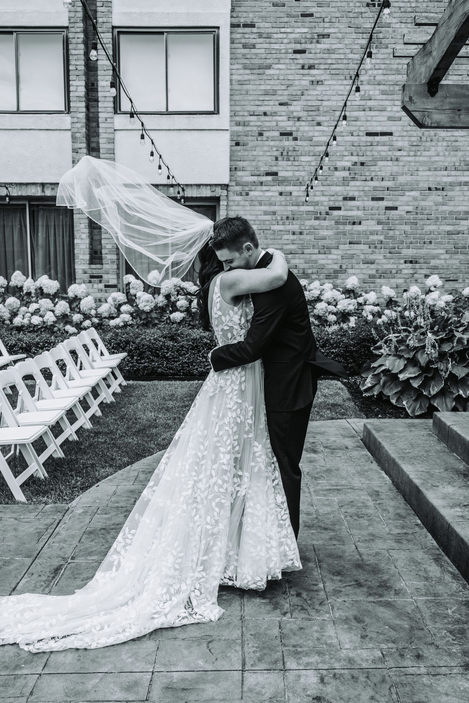 Black and white photo of newlyweds embracing on patio with flowing veil and string lights.