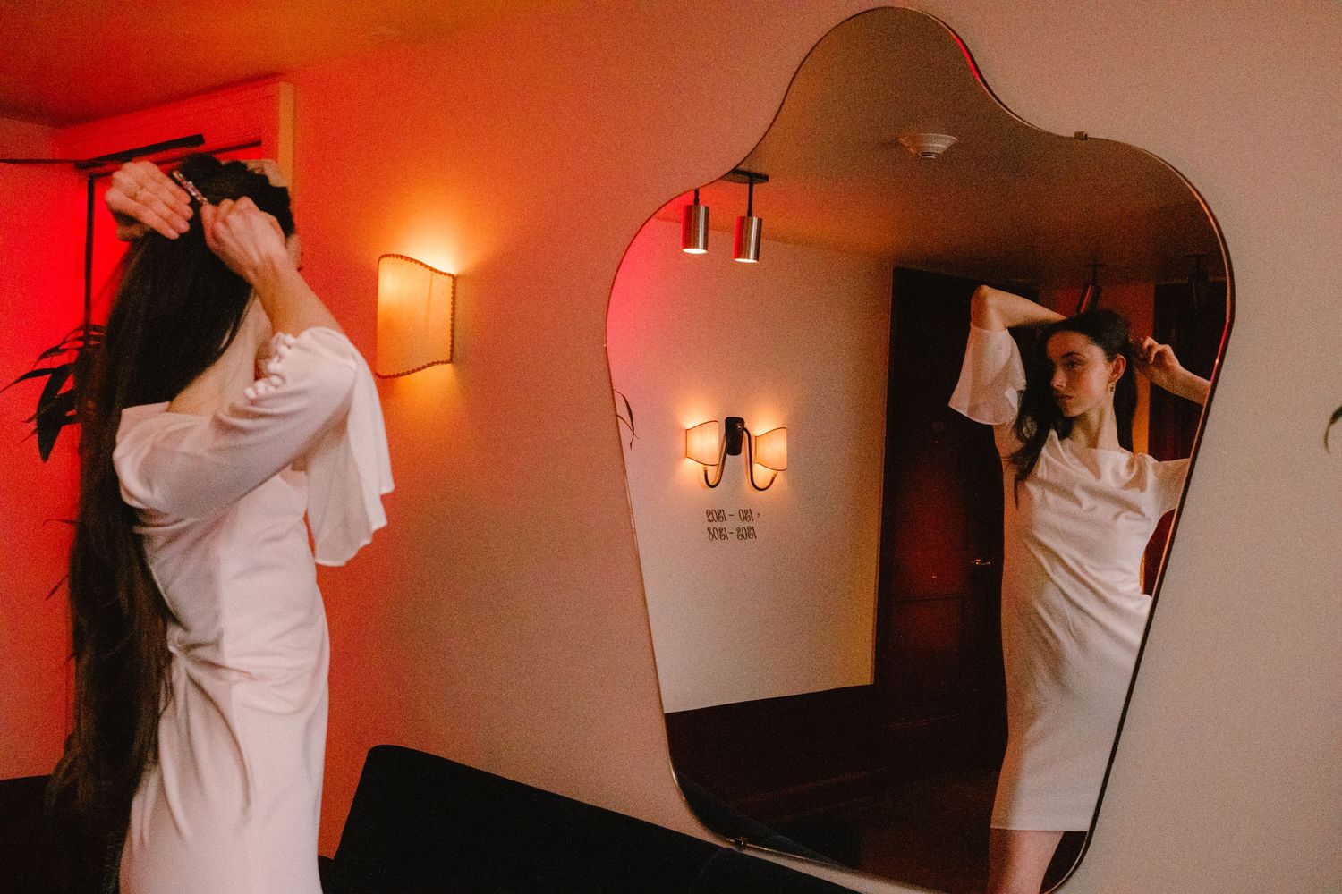 Woman in white robe adjusts her long dark hair while standing before a large asymmetric mirror in warm-lit room.